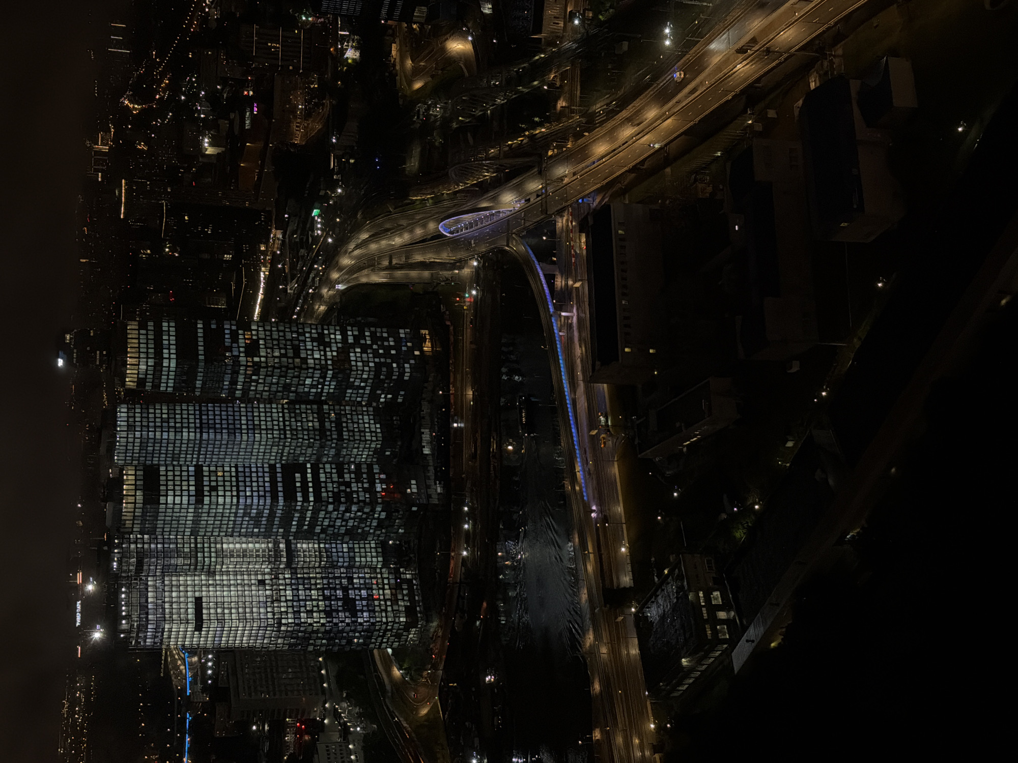 This aerial, wide-angle photograph captures a nighttime cityscape in Khimki, Russia, focusing on the Moscow International Business Center (MIBC) complex, also known as Moscow City. The dominant features are several illuminated skyscrapers with grid-like patterns of windows, reflecting a cool blue and white light. In the foreground and midground, a multi-lane highway curves through the scene, its lanes marked by bright streetlights and the headlights and taillights of a few vehicles. A distinctive arched bridge, illuminated with blue lights, spans across the highway. Below the highway and to the left, a dark body of water, likely the Moskva River, shows ripples and reflections from the surrounding lights. Several lower-rise industrial or commercial buildings with dimly lit windows are scattered throughout the lower portion of the frame, contrasting with the towering skyscrapers. The distant background reveals a dense urban sprawl of more illuminated buildings and faint lights extending towards the horizon under a dark, hazy sky. The overall scene is one of a dynamic, modern city at night, characterized by artificial illumination and extensive infrastructure.