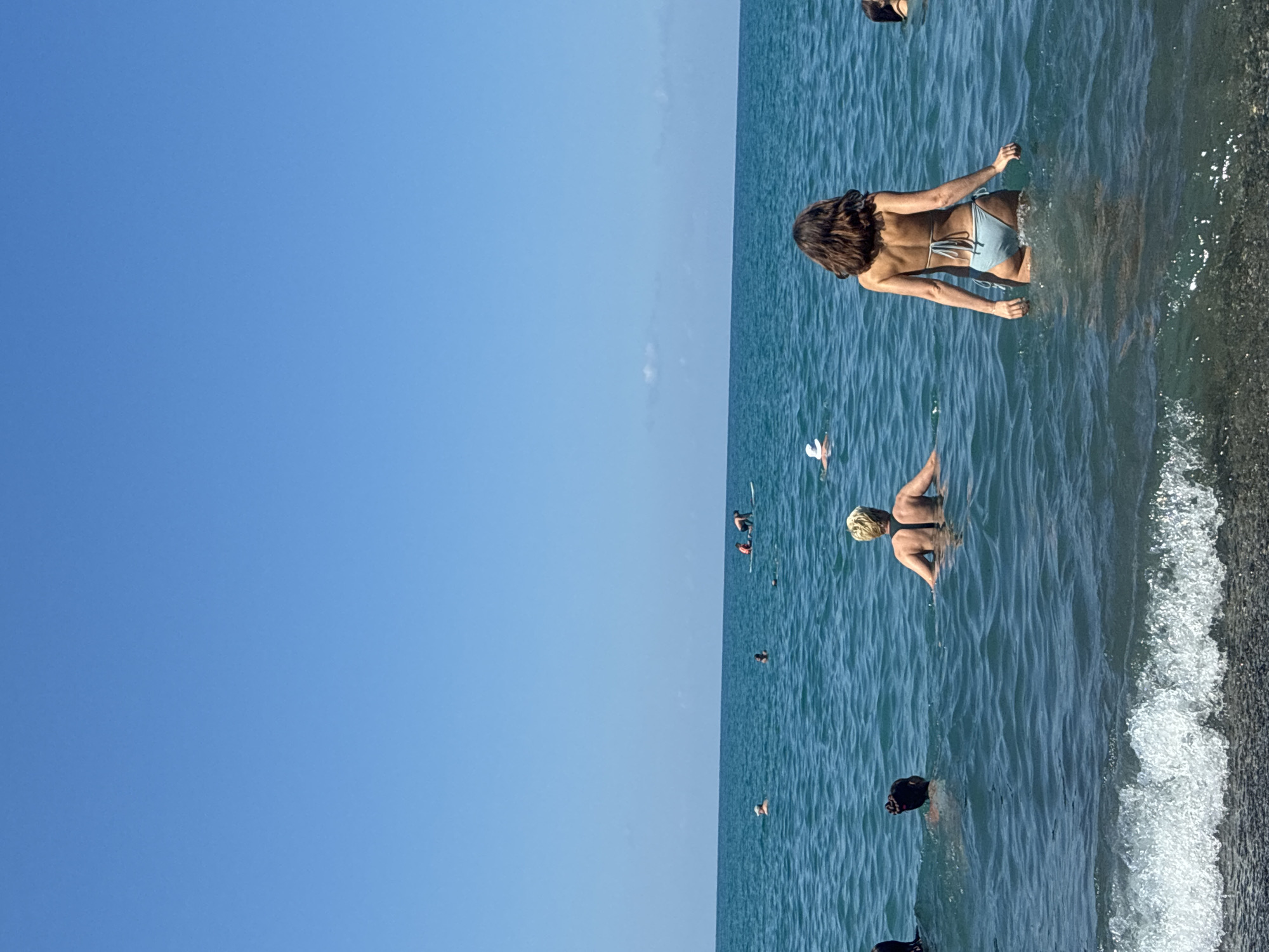 This is a medium shot, taken from a low angle, of people in the sea on a sunny day. The sky is a clear, bright blue. The sea is a deep blue-green, with gentle waves. In the foreground, the edge of the sea meets a pebble beach.
There are several people in the water. In the middle ground, a woman with blonde hair is facing away from the camera, with her arms outstretched. To her right, a woman with dark brown hair is also facing away from the camera, wearing a light blue bikini. Further in the distance, there are a few other individuals and a paddleboarder with two people on it, one of whom appears to be standing.
The scene is likely the Black Sea coast in Sochi, Russia, a popular resort city known for its beaches and outdoor activities.