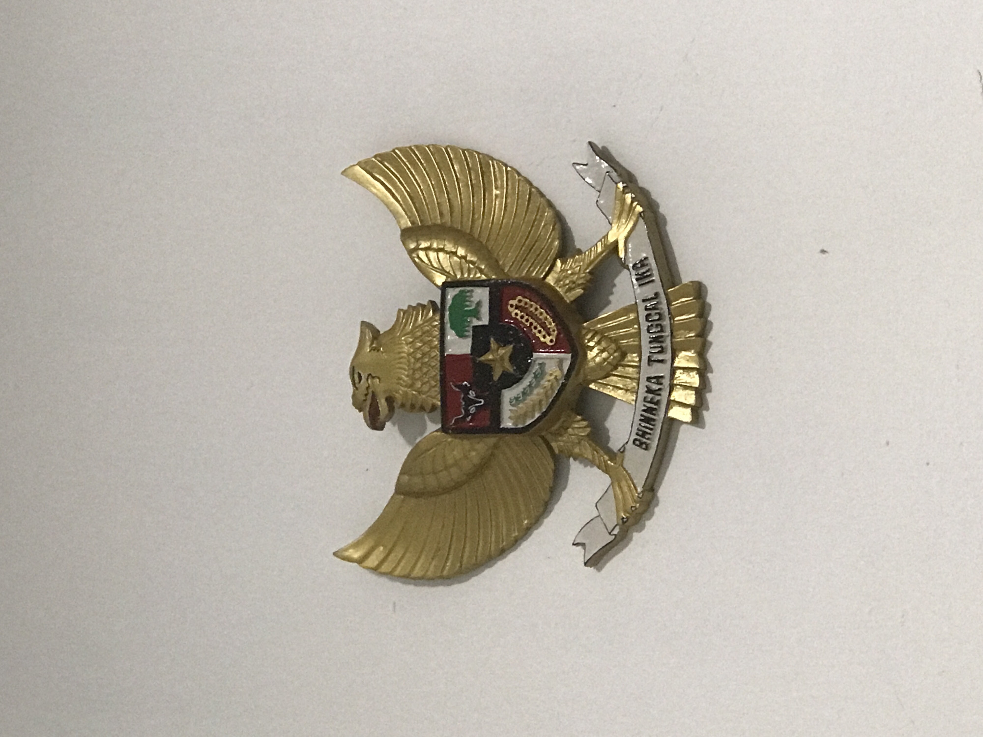 A close-up shot shows a golden Garuda Pancasila emblem, the national symbol of Indonesia. The emblem is against a plain, light-colored background. The Garuda (a mythical bird) has its wings spread, and a shield divided into five sections is on its chest. The five sections represent the five principles of Pancasila, Indonesia's state philosophy. A banner reading "BHINNEKA TUNGGAL IKA" (Unity in Diversity) is clutched in the Garuda's talons. The scene is not located in the city of Lhokseumawe, Indonesia, as there is no geographical evidence to support that.