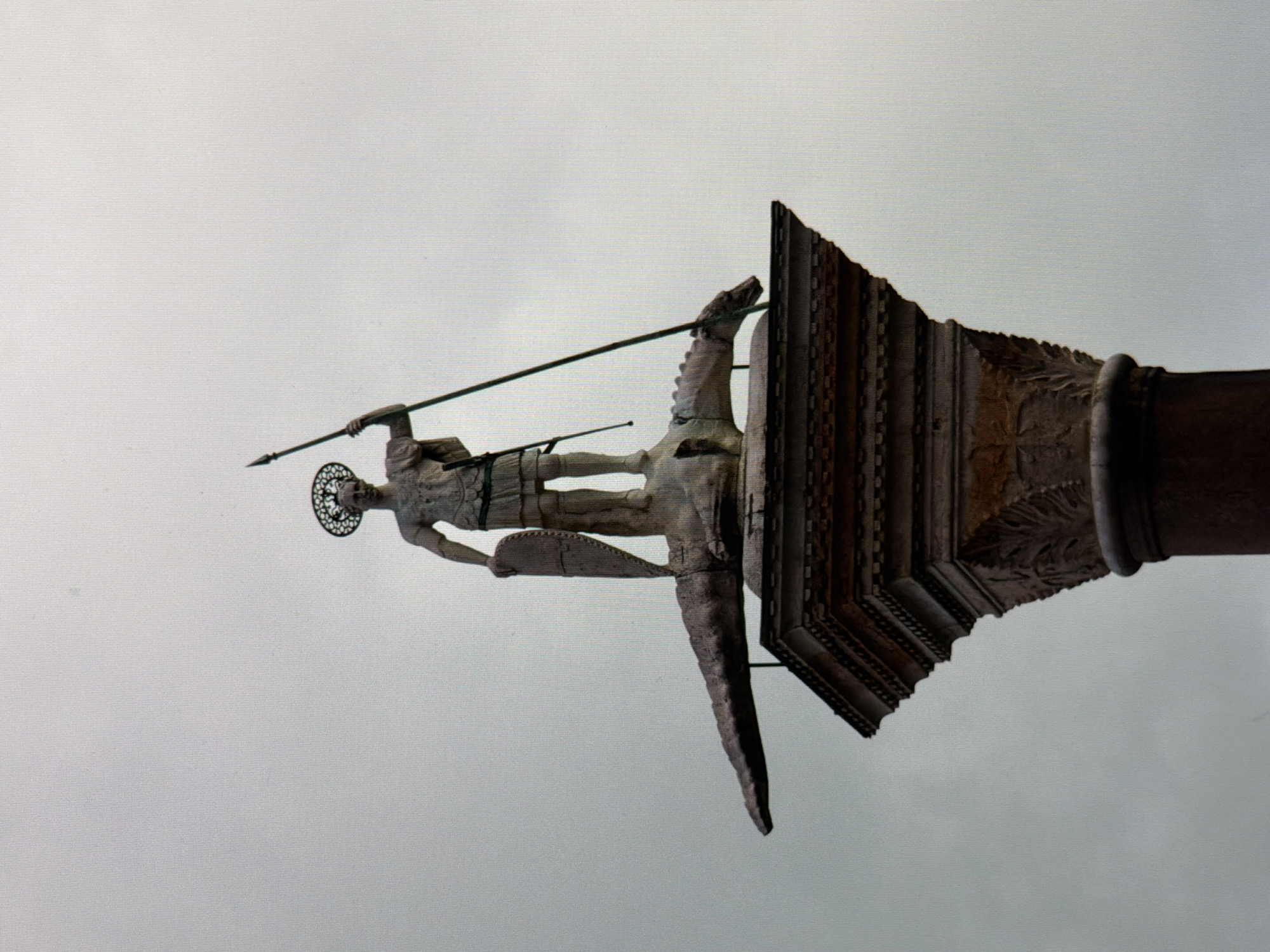 The image shows the statue of St. George, the dragon-slayer, atop a column in St. George's Square in Kyiv, Ukraine. The bronze statue depicts St. George, a soldier with a halo, standing over a slain dragon. He holds a spear in his right hand and a shield in his left. The statue is weathered and shows signs of age. The column itself is ornate, with carved details. The sky is overcast and gray, suggesting a cloudy day. The background is plain, with no other notable features. The image is taken from a low angle, looking up at the statue.