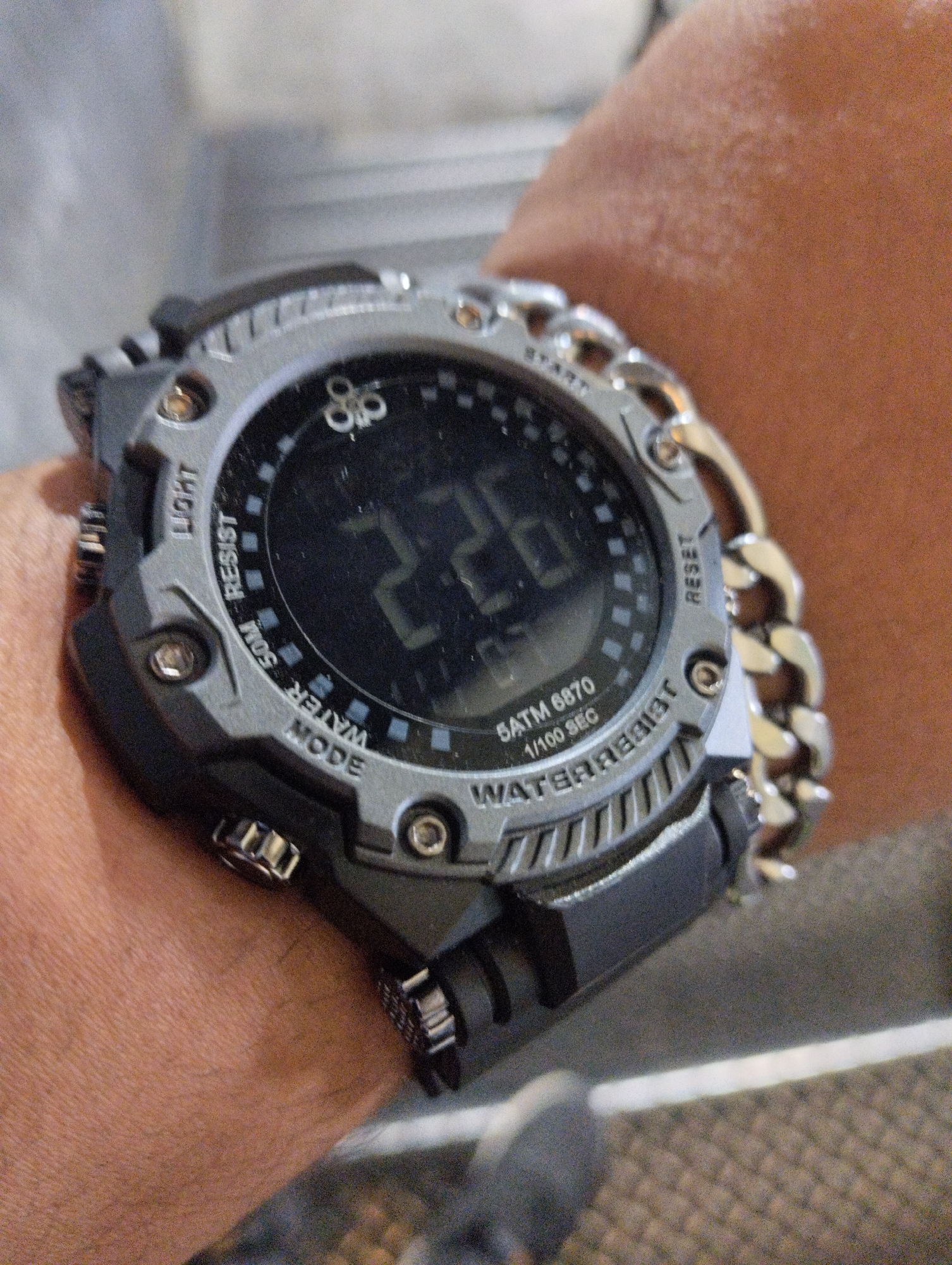 A close-up photograph shows a digital watch on a person's wrist. The watch face displays "2:26". The watch has "LIGHT", "RESIST", "MODE", "START", and "RESET" printed on it. A silver chain acts as the watch band. The scene is indoors and lacks discernible contextual city or environmental details for identification.