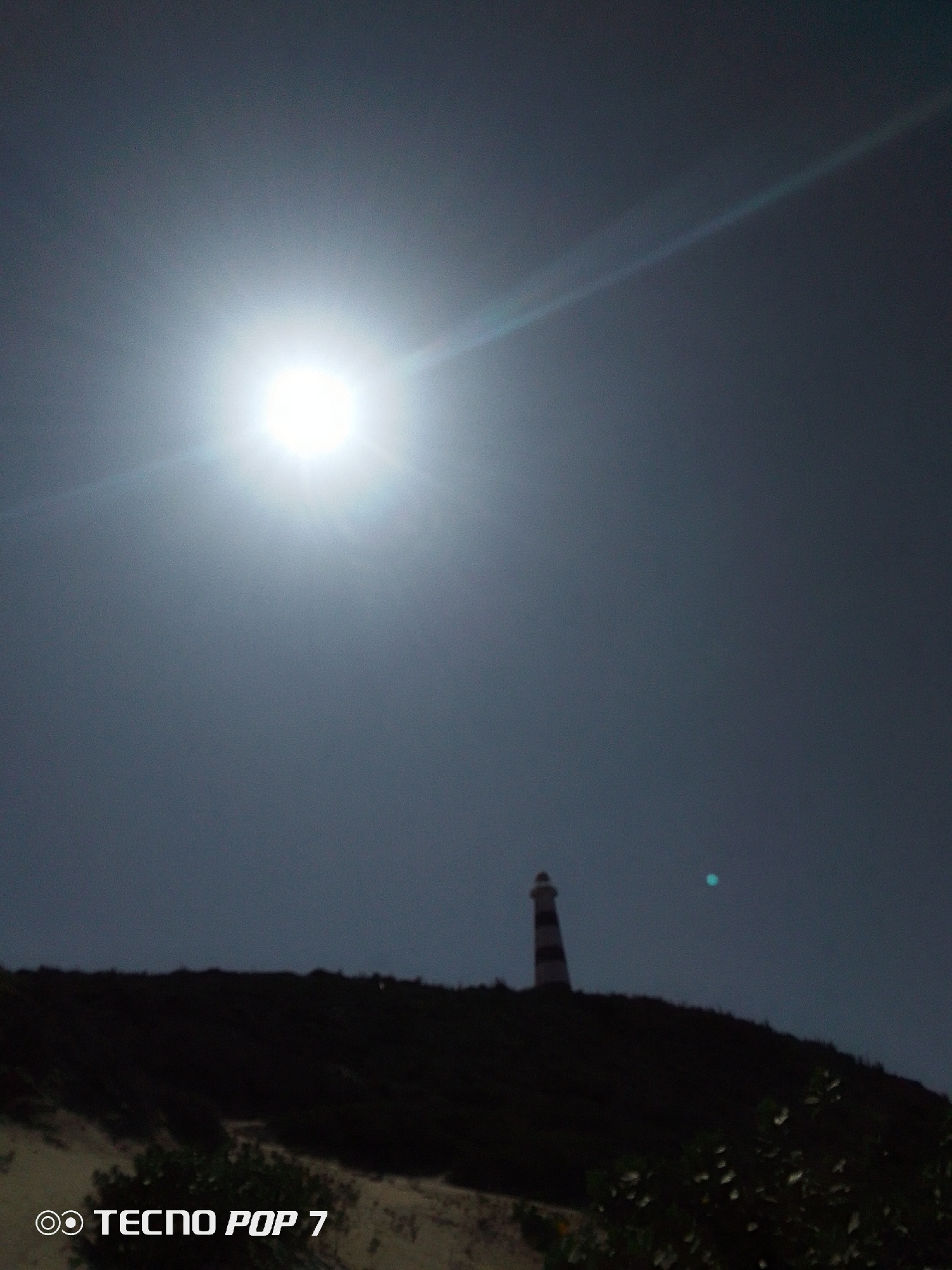 The image shows a lighthouse on top of a hill, silhouetted against a bright, overcast sky with the sun visible at the top of the frame. The location is an unknown city and location, as no identifying information is present. The hillside is dark and indistinct, with some vegetation visible in the foreground. There are no people visible in the image. A bright spot, possibly a lens flare, is visible in the sky to the right of the lighthouse. The words "Tecno Pop 7" appear at the bottom left corner of the image.