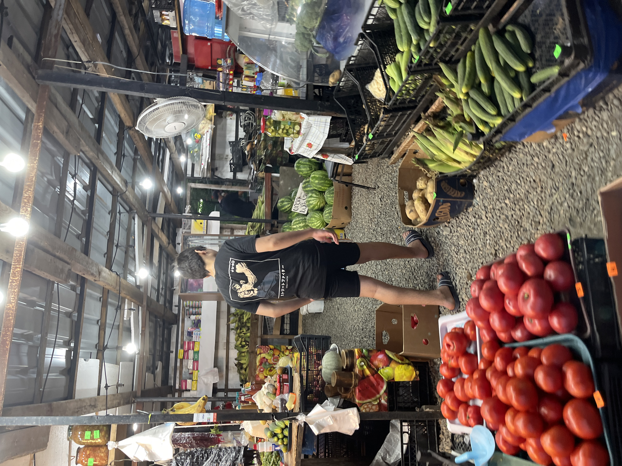 A young male, appearing to be in his late teens or early twenties, stands with his back to the camera in a bustling outdoor market stall in Ureki, Georgia. He is wearing a black t-shirt with a graphic print of two figures in a martial arts pose and black shorts, with blue and white flip-flops on his feet. The stall is filled with a variety of fresh produce, including large piles of ripe tomatoes in trays, watermelons, cucumbers, corn, and potatoes in boxes. Jars of pickles and other preserved items are visible on shelves and tables. Overhead, a fan is mounted on a post, and bare light bulbs illuminate the market, suggesting it is either dusk or an indoor/covered area. The ground is covered in gravel. In the background, other market-goers and more stalls can be seen, indicating a lively commercial environment. The overall scene depicts a typical day at a local Georgian produce market.