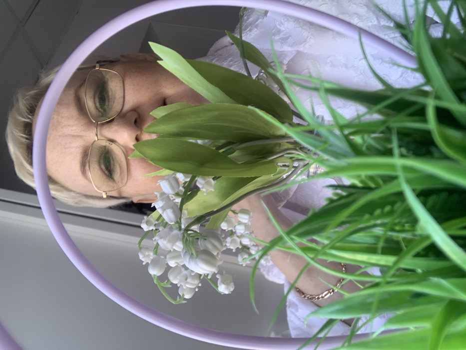 The image depicts a close-up, low-angle view of an adult female partially obscured by artificial flora and a decorative object in an indoor environment. In the immediate foreground, a mass of out-of-focus artificial green grass occupies the bottom third of the frame. Above the grass, a bouquet of artificial white lily-of-the-valley flowers with prominent green leaves is centered. A segment of a lavender-colored hoop is visible to the left of the flowers, curving upwards through the midground. The upper portion of a female's face is visible behind and above the bouquet; she has short, blonde hair and wears gold-rimmed glasses, looking directly forward. Her right arm, adorned with a gold link bracelet, is partially visible to the left of the bouquet. She is wearing a light-colored, textured garment, possibly a white blouse. The background consists of a uniformly light-colored wall or window, indicating an indoor setting. The specific location within the city of Chaikovsky, Russia, is not discernible from the visual information.