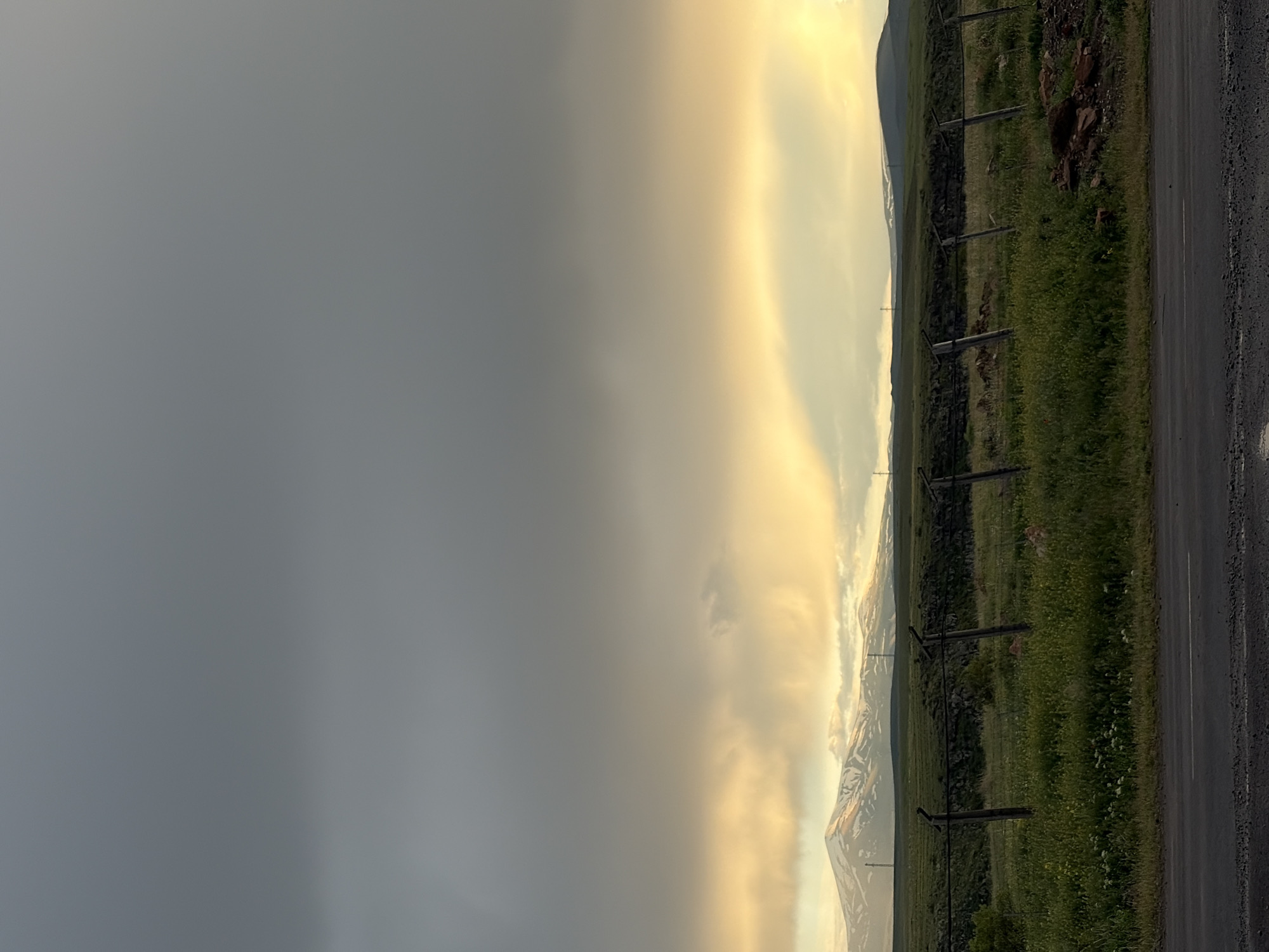 The image shows a landscape in Anipemza, Armenia. On the right side is a road with vegetation and a fence line. In the distance, mountains are visible, partially obscured by clouds and what appears to be a weather front or cloud formation. The sky is divided between a dark, overcast area on the left and a lighter, brighter area near the mountains.