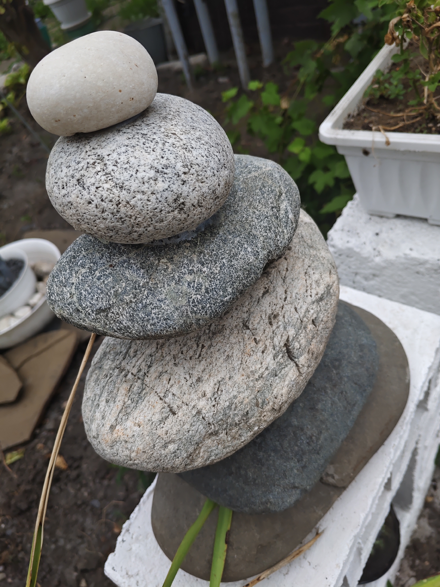 A close-up, high-angle shot captures a stack of smooth, rounded river stones precariously balanced on top of one another. The stones vary in size and color, ranging from a light off-white on top to darker grays and speckled granite tones lower down. The stack is situated outdoors, with a blurred background suggesting a garden setting. Green foliage and a white planter are visible to the right, and a light-colored wooden object and dark soil are in the lower left. A thin, dry reed or grass stalk leans against the stone stack. The lighting is natural, indicating daytime. The scene appears to be located in a backyard or garden area within Rostov-on-Don, Russia, possibly as part of a decorative arrangement or an attempt at stone balancing art. No people are present in the image.