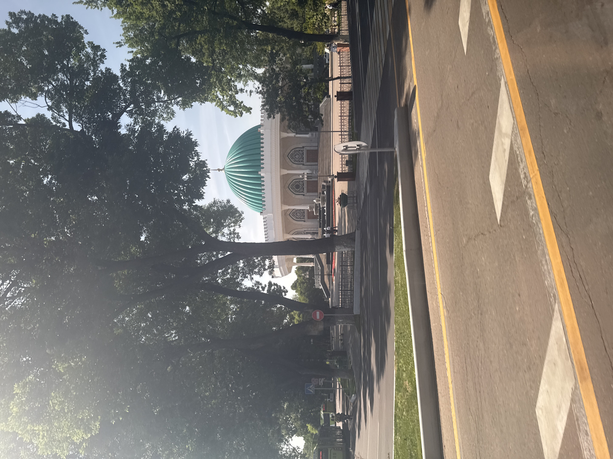 The image depicts a mosque with a prominent turquoise dome, situated in Kashgar mahalla, Uzbekistan. The mosque is surrounded by mature trees, partially obscuring the view. A street runs alongside the mosque with white and yellow lane markings. There are no people in the foreground. A fence surrounds the mosque. Signage is visible.