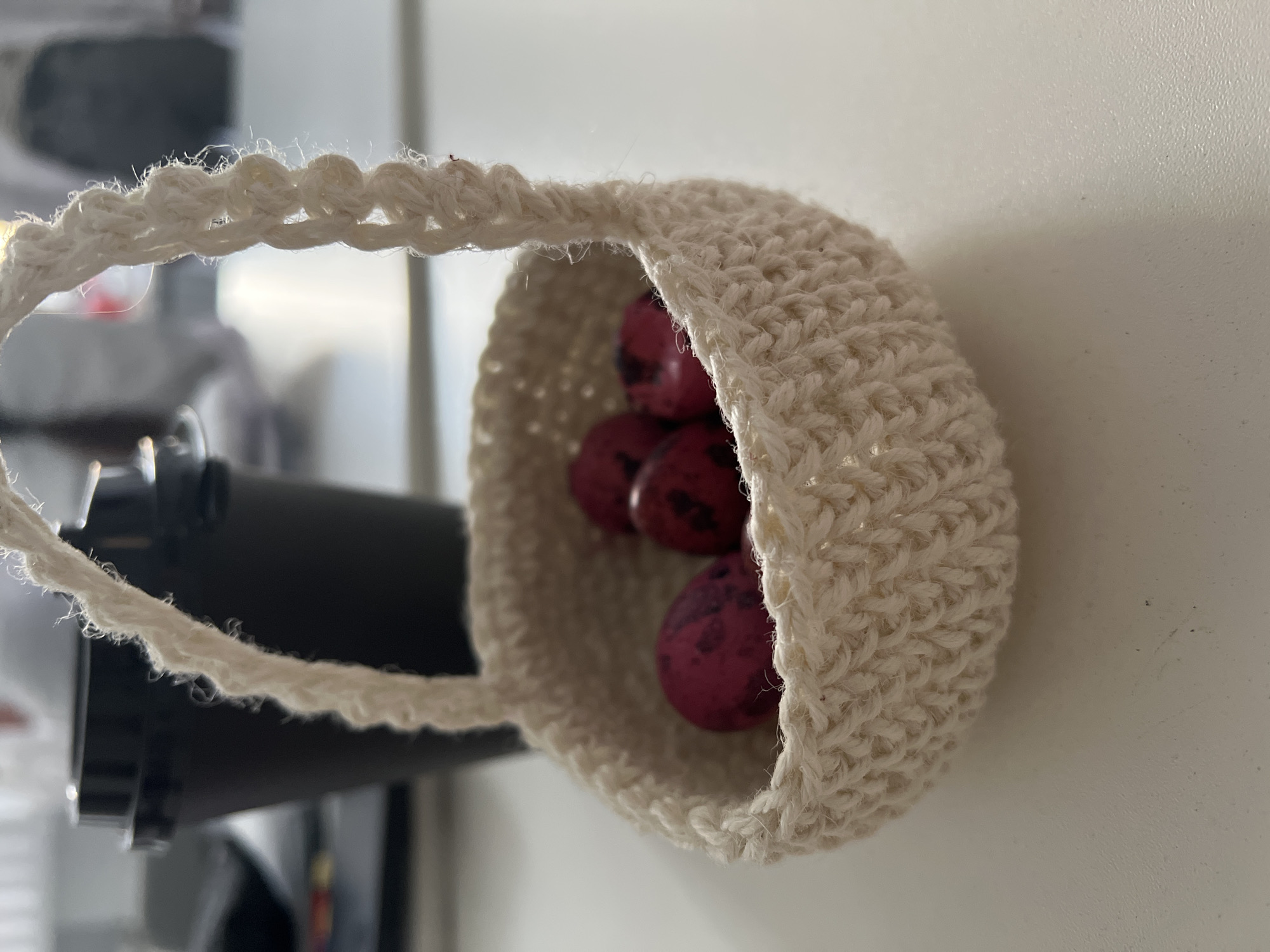A close-up, slightly tilted, shot features a small, crocheted, off-white basket filled with speckled, reddish-purple eggs. The basket has a crocheted handle. In the blurred background, a dark object, possibly a piece of equipment, is visible. The background also includes a white surface, possibly a wall. The scene does not show Moscow, Russia.