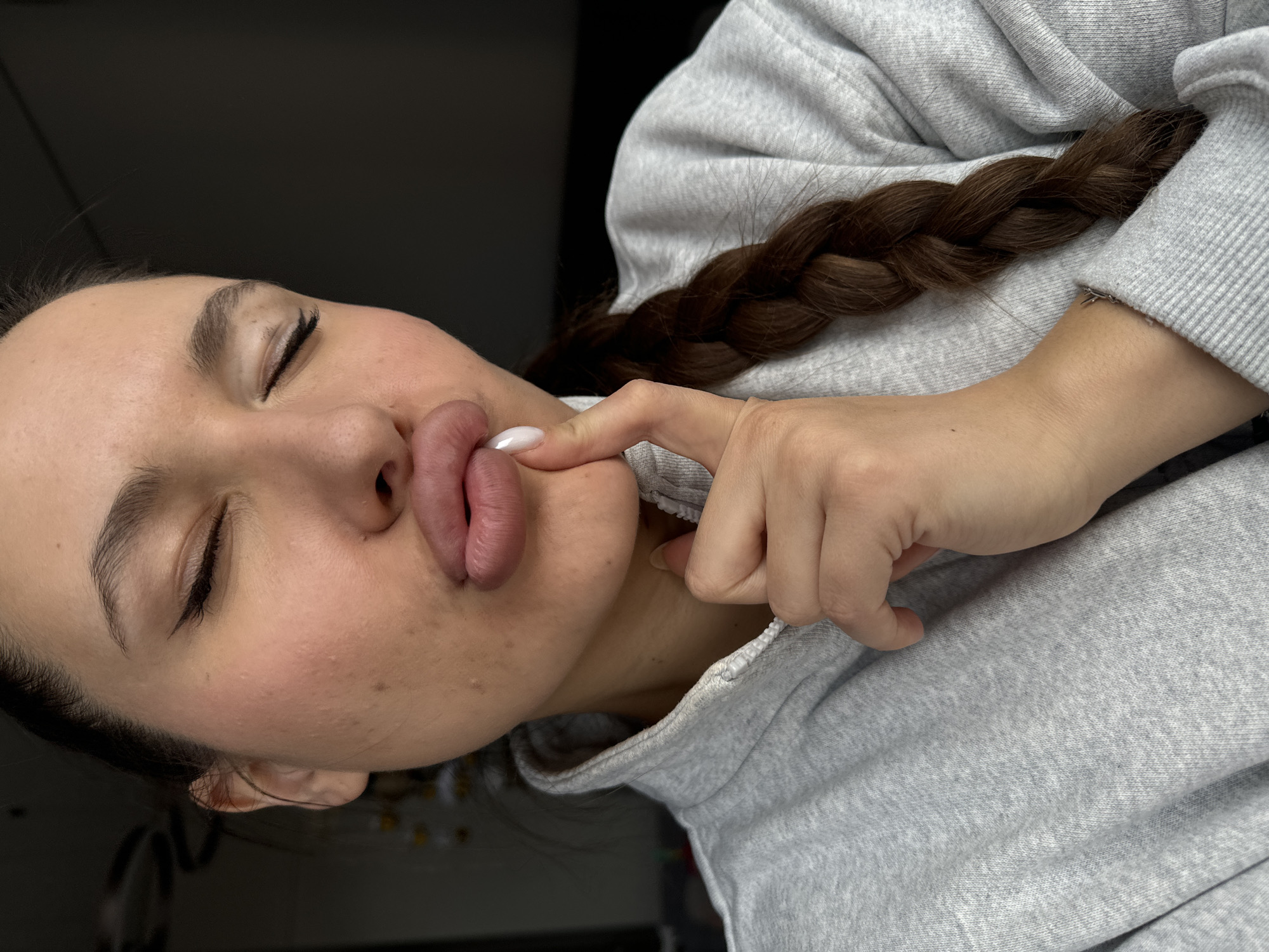 A woman with closed eyes and pursed lips is photographed indoors. Her skin appears fair, with some visible blemishes on her cheeks. She is wearing a gray hooded sweatshirt. Her brown hair is styled in a braid that lies over her shoulder. Her right hand is visible, with a finger touching her chin. Her fingernails appear to be manicured with light-colored polish. The background appears to be a dark or dimly lit area. There is no information on the location within Odintsovo, Russia in the scene.