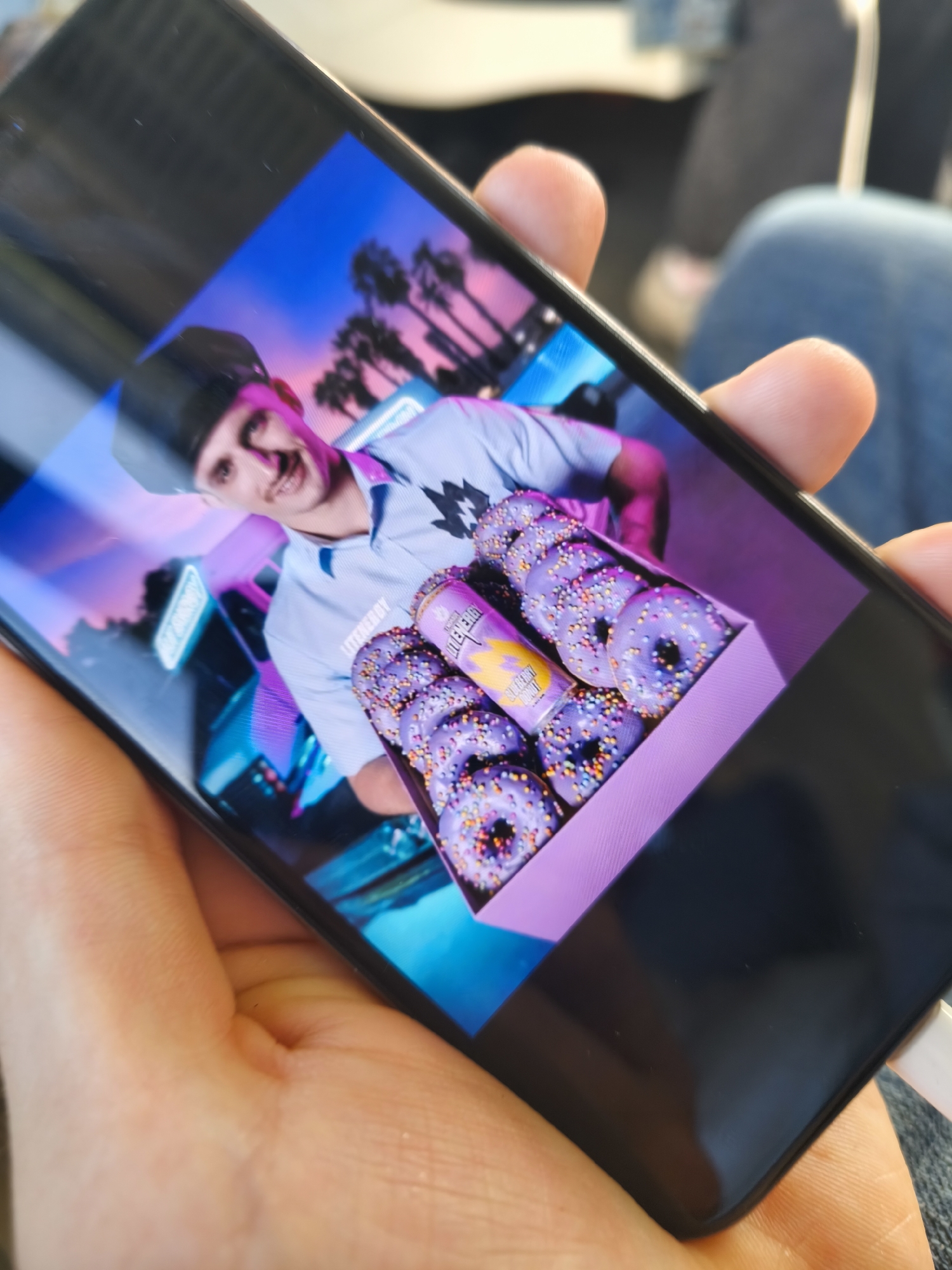A smartphone displays an image of a person wearing a light blue collared shirt and a black cap, smiling at the camera. The person is holding a box of purple donuts with sprinkles and a can of a purple drink. The background of the image is a neon-lit city scene with palm trees. The phone is held by a hand with visible fingers. The image is blurry and appears to be taken at an angle.