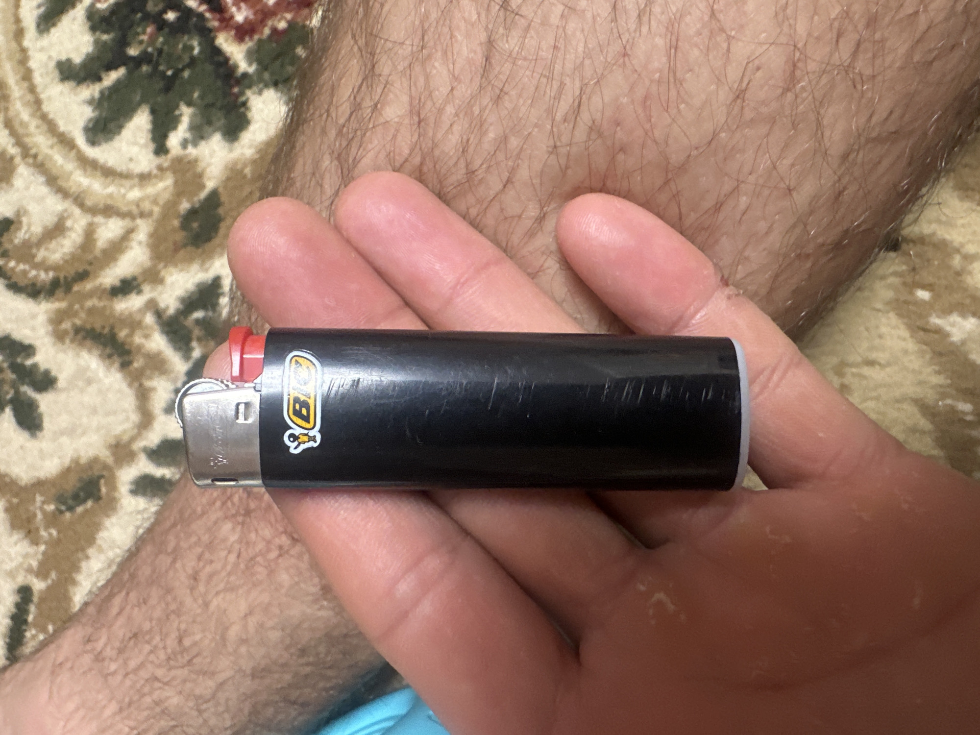 The image shows a hand holding a black BIC lighter. The lighter has a silver metallic top with a red plastic ignition wheel. A yellow BIC logo with a cartoon figure is visible on the side of the lighter. The hand appears to be that of a person with dry skin. The background consists of a fuzzy carpet with a patterned design in shades of brown, beige, and green. The lighting suggests an indoor environment. There is no discernible city or location information present in the image.