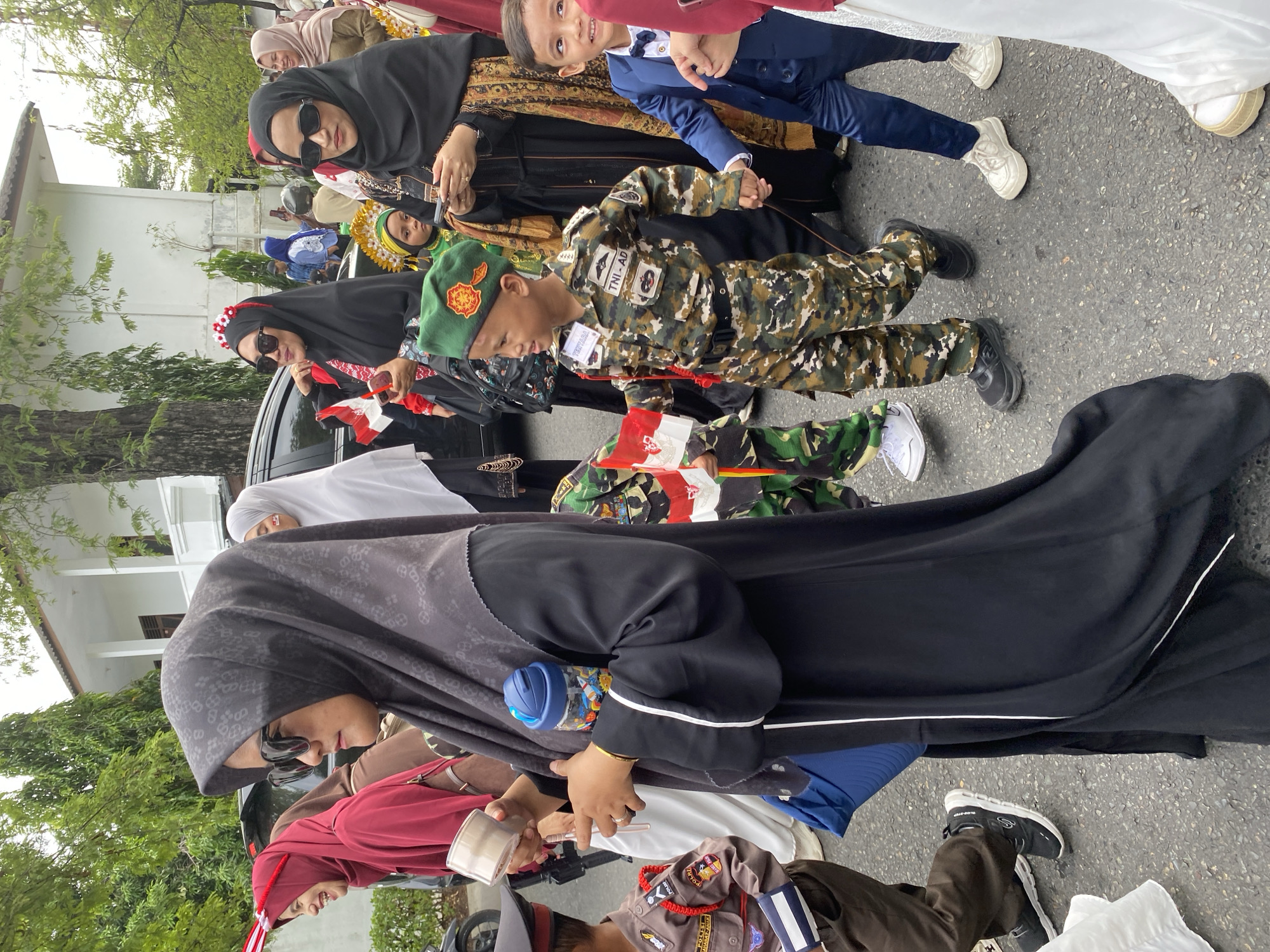 In an outdoor scene in Banda Aceh, Indonesia, a procession is underway. The foreground is dominated by a woman wearing a black hijab and sunglasses, holding a blue water bottle filled with small toys and a beige cylindrical object. To her left, a young boy in a camouflage military uniform, complete with a green beret, walks forward, holding a small Indonesian flag. Beside him, another young boy in a blue suit and bow tie is being led by an unseen adult. Several other individuals are visible in the background, including women in hijabs and children. The environment appears to be a street with trees and a white building in the background.