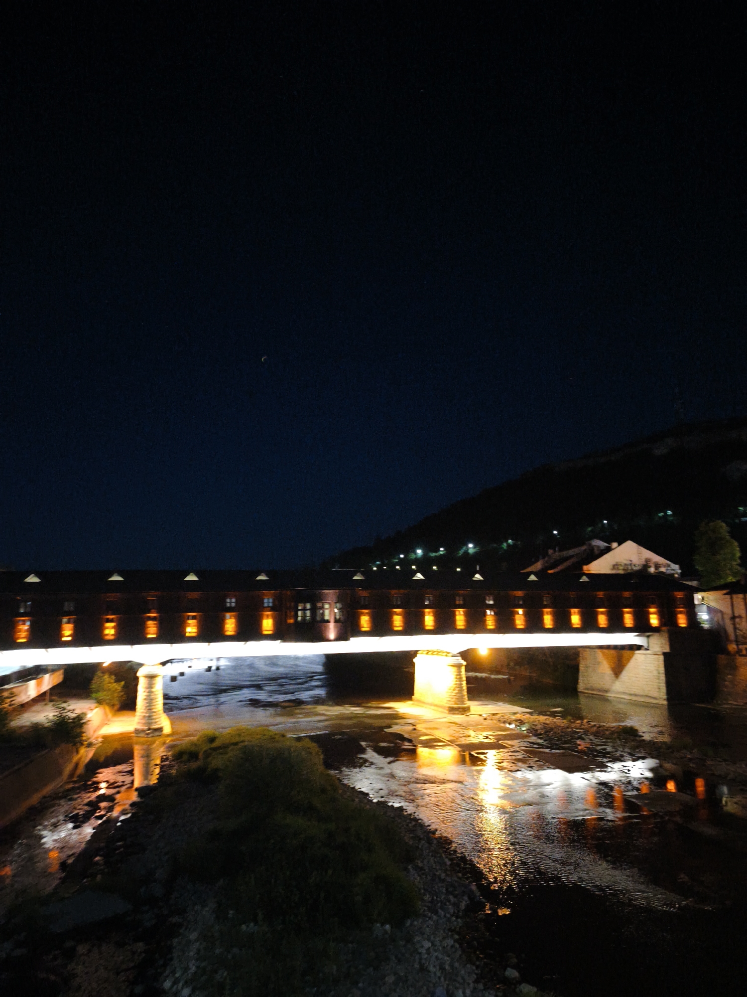 This is a nighttime photograph of the covered bridge in Lovech, Bulgaria. The bridge, which spans a river, is illuminated by a series of warm, yellow lights. The lights reflect on the surface of the water, creating a shimmering effect. The bridge is a long, rectangular structure with numerous windows, each with an individual light source, giving the impression of a continuous row of lit rooms. A hill with scattered lights can be seen in the background, suggesting a settlement or town. The sky above is dark, with a faint crescent moon visible. The foreground shows the riverbank with some vegetation and rocks.