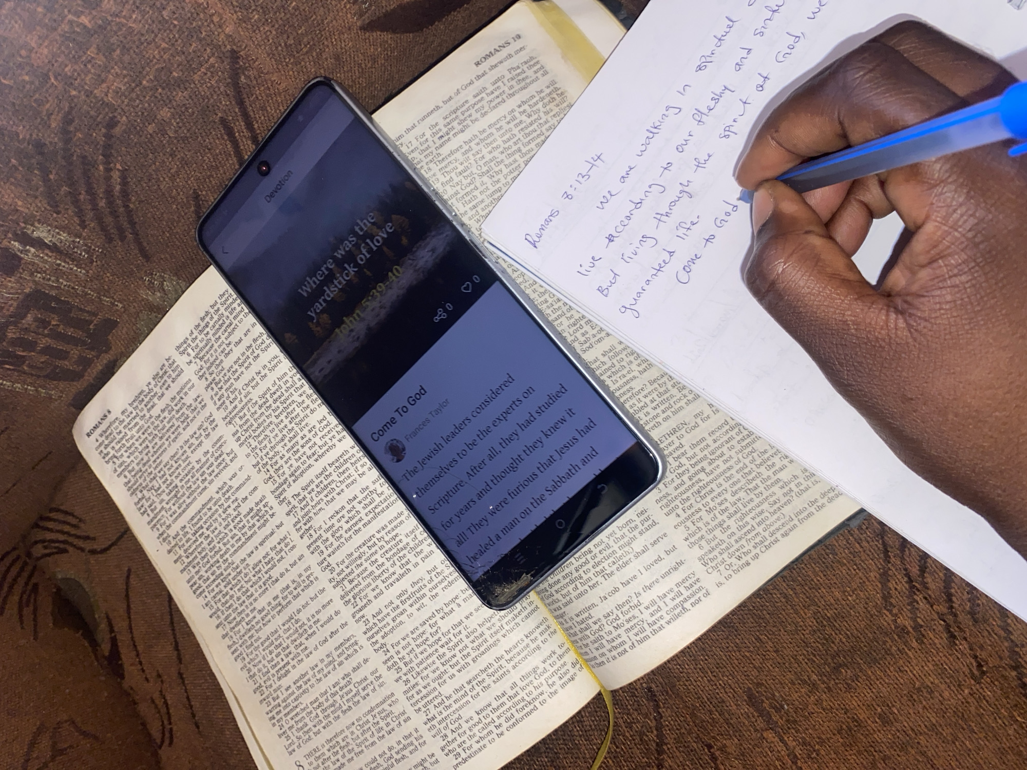 The image captures a personal moment of spiritual study, featuring a dark-skinned hand writing notes in a white notebook, an open Bible, and a smartphone displaying a devotional app, all resting on a dark, textured surface, possibly wood or fabric.
In the foreground to the right, a dark-skinned hand holds a blue ballpoint pen, actively writing on a white sheet of paper. The handwriting, in blue ink, is cursive and includes several lines of text. Partially visible notes read: "Romans 8:13-14", "we are walking in spiritual", "live according to our fleshy and sinful", "But living through the spirit of God, we", "guaranteed life.", and "Come to God."
To the left of the hand and notebook, a black smartphone with a narrow bezel is visible, resting diagonally across an open Bible. The phone's screen is on, displaying content from a devotional application. At the top of the screen, the word "Devotion" is visible, along with a back arrow icon. The main title of the devotional is "Where was the yardstick of love", with a subtitle "John 5:39-40". Further down, the author is identified as "Frances Taylor", under the heading "Come To God". A snippet of the devotional text is also visible: "The Jewish leaders considered themselves to be the experts on Scripture. After all, they had studied for years and thought they knew it all! They were furious that Jesus had healed a man on the Sabbath and". The bottom right corner of the phone screen shows a small crack.
Beneath the smartphone and partially obscured by it, an open Bible lies flat. The visible pages show text in a two-column format with small verse numbers, characteristic of a Bible. Headings "ROMANS 8" and "ROMANS 10" are clearly identifiable on the left and right pages, respectively. The text on the pages includes verses from Romans, such as "For to be carnally minded" and sections discussing being "dead to the law" and "living in the Spirit". A yellow ribbon bookmark is visible near the spine of the Bible, peeking out from under the phone.
The entire arrangement is set against a dark brown, textured background, which appears to be a table surface or a dark, woven fabric. The lighting suggests an indoor setting, possibly with an overhead light source illuminating the scene.
