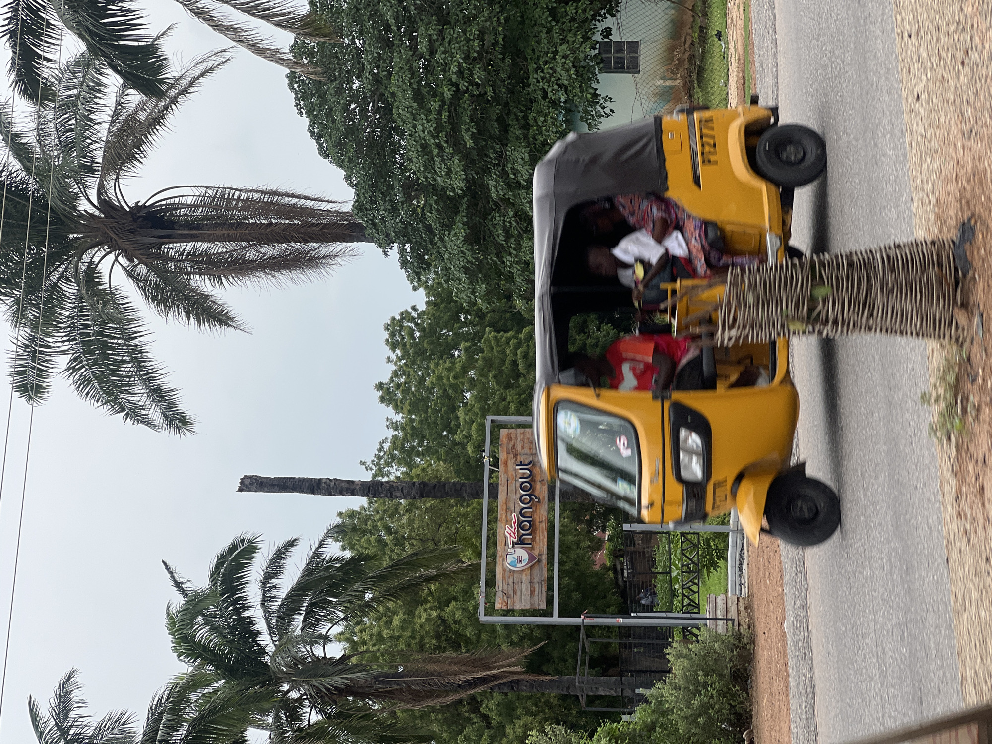 In Jalingo, Nigeria, a bright yellow auto-rickshaw, a common form of transportation in the region, travels along a paved road under an overcast sky. The auto-rickshaw carries four occupants: a man in a red shirt driving, a man in a white shirt, and two women. The auto-rickshaw has a black canvas roof and a license plate that reads "FG277R". To the left of the road, a wooden sign with the words "the hangout" in a stylized font is visible, flanked by palm trees and lush green foliage. In the background, a light blue building with a window and a chain-link fence can be seen. The scene is characterized by a tropical environment, with tall palm trees and dense greenery dominating the landscape.