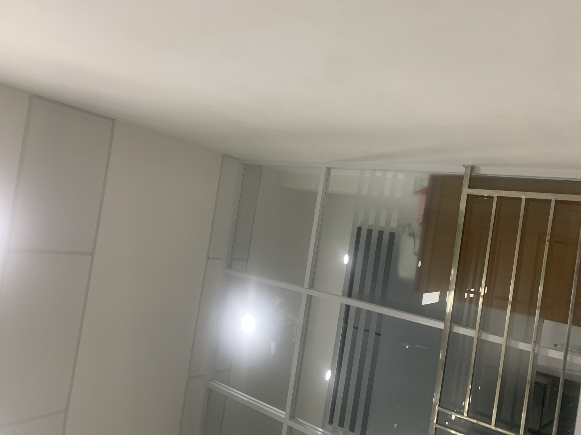 The image shows an interior scene, possibly an office or commercial space, with a white tiled ceiling. There are glass partitions with white metal frames, creating distinct sections within the space. A bright overhead light is visible, reflecting off the glass. Below the glass partitions, there is a dark wooden cabinet or counter with a reflective surface. In the foreground, a metallic railing with horizontal bars suggests a balcony or elevated walkway. The walls are painted white, and the overall impression is one of a modern, clean, and minimalist design. The image does not contain any explicit visual cues that would allow for specific location identification within Palembang, Indonesia.