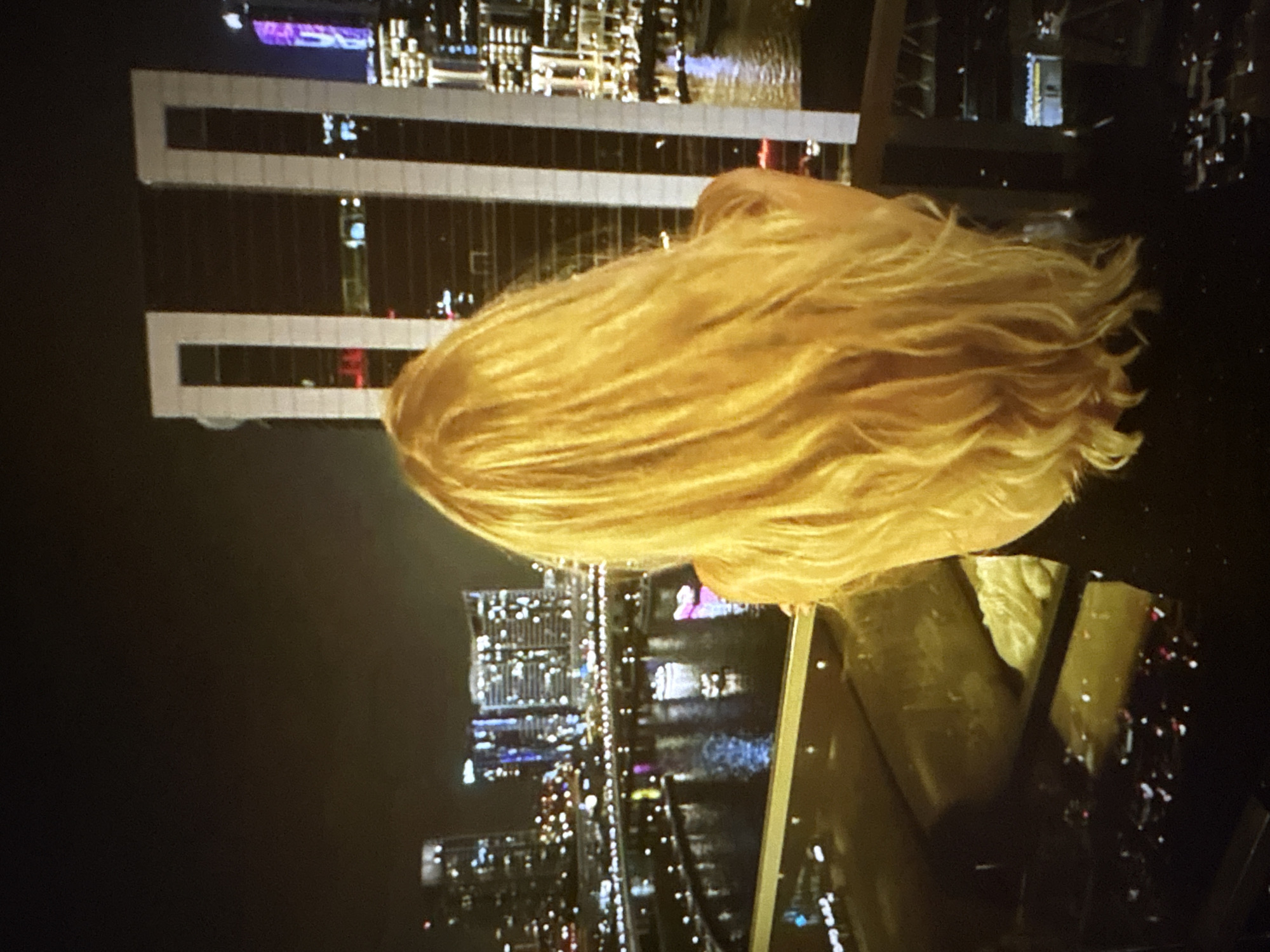 The image shows a woman from behind with long blonde hair, standing on a balcony overlooking a city skyline at night. The city is illuminated by numerous lights from buildings and streetlights, and a body of water reflects the lights. A distinctive building with large rectangular cutouts is visible in the background. The scene is dark, with the primary light sources being the city lights and the artificial lighting on the woman. The woman is wearing a dark top. The location is not Pori, Finland, but rather a modern city with skyscrapers and a waterfront.