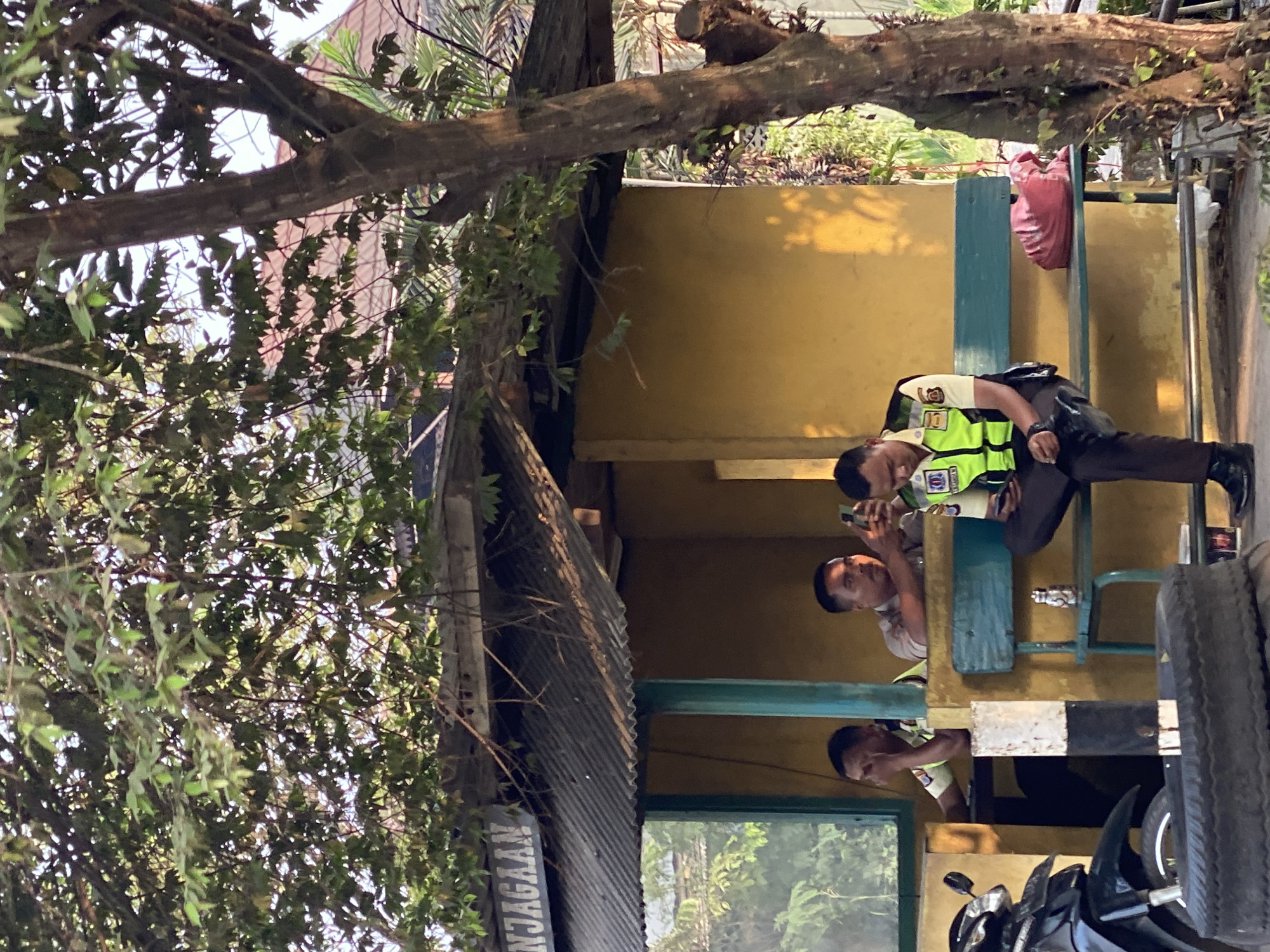 In the city of Kasihan, Indonesia, an outdoor scene depicts a guard post with three individuals. Two are seated on a bench, one on the right wearing a reflective vest over a light-colored uniform, engrossed in their smartphone. To their left, another individual in a similar uniform leans on the bench, also looking at a phone. The third person, to the far left and partially obscured, is also in uniform and seated. A motorcycle is parked to the left of the post, with a spare tire in the foreground. The guard post itself has a corrugated metal roof and a yellow-painted wall, with a sign reading "NJAGAAAN" above the entrance. Lush green foliage from overhead trees creates a dappled light effect on the scene.