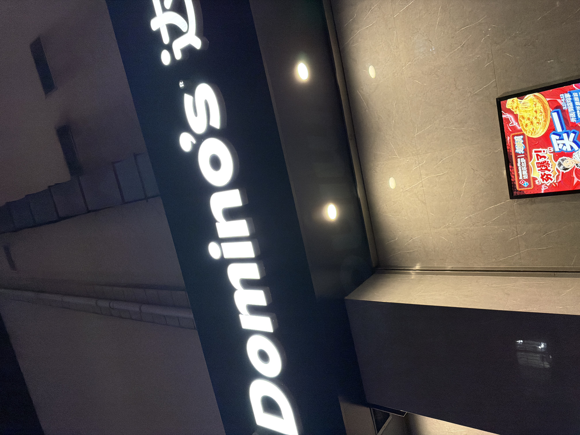 The image presents a nighttime, angled view of a Domino's Pizza storefront located in Jiangning District, China. The upper portion of the frame features a prominently illuminated sign displaying the white English lettering "Domino's®," followed by the partial Chinese characters "达" (Dá). Below this primary signage, a dark grey soffit with two circular recessed spotlights emitting warm light is visible. The building's facade beneath the soffit is faced with light-colored, patterned stone or tile, also illuminated by additional recessed lighting fixtures. In the lower right quadrant of the image, a rectangular, red-framed promotional display or digital screen is positioned, showcasing a graphic of a pizza with melted cheese, Chinese promotional text including "买一" (Buy One), and a cartoon character. No individuals or specific actions are discernible within the frame.