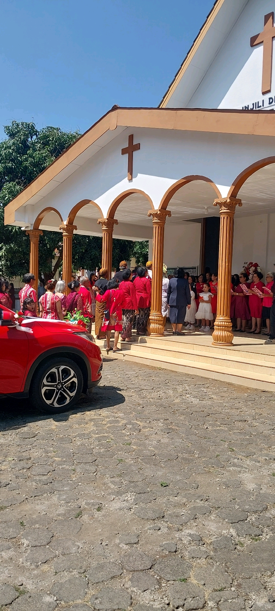 In Keling, Indonesia, a group of people, many dressed in traditional red and gold attire, gather outside a church. The church features a white facade with a brown-trimmed roof and arched entrances supported by ornate columns. Two large brown crosses are visible on the church's exterior, one on the peak of the roof and another above the main entrance. A red car is parked to the left of the frame, decorated with white bows and a bouquet of flowers. The ground is paved with hexagonal stones, and trees are visible in the background under a clear blue sky. The scene appears to be a celebratory event or gathering outside a place of worship.