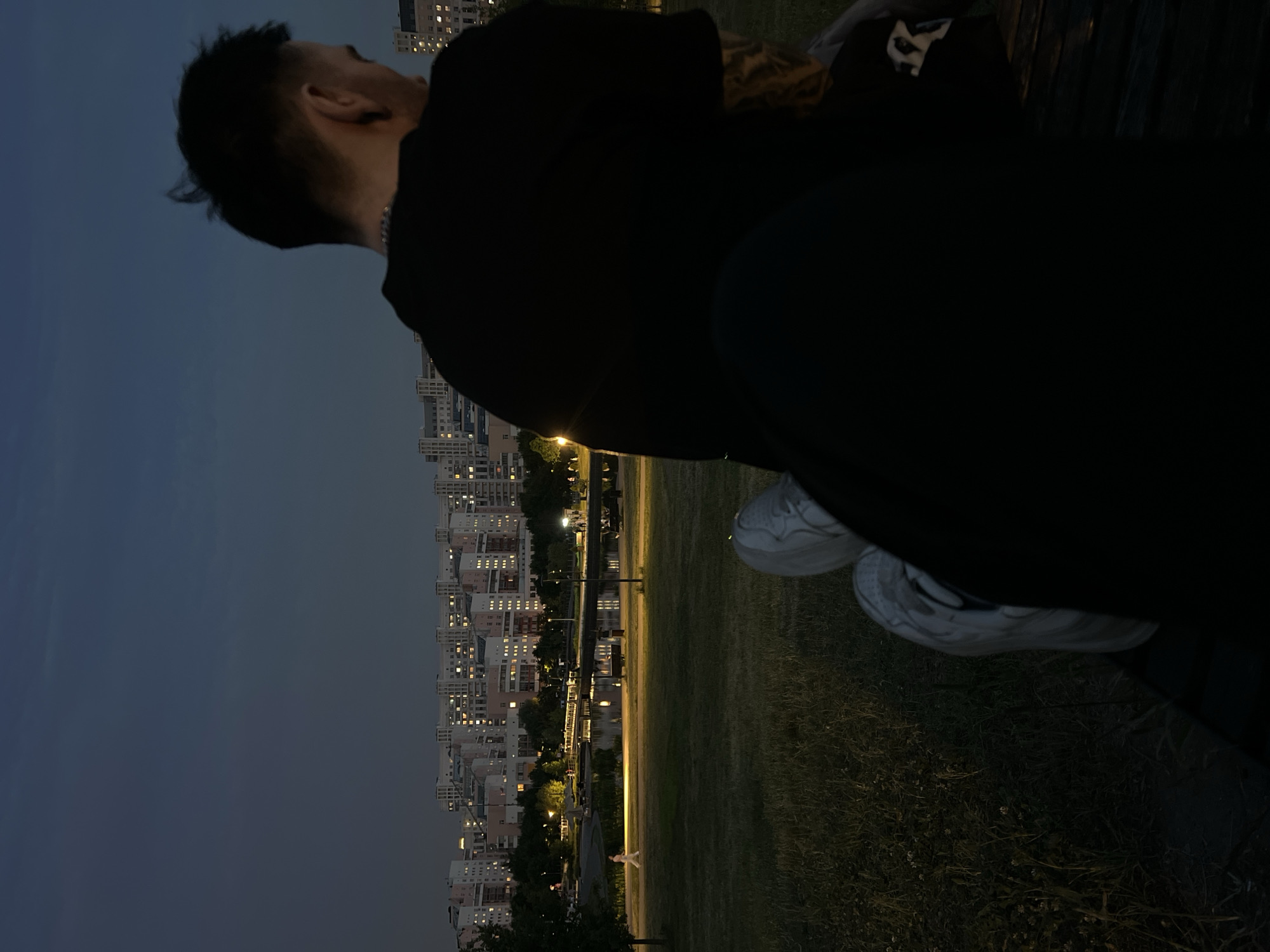 The image shows a night scene in a park in Khimki, Russia. In the foreground, a person is sitting on a bench, their back to the camera, wearing a black t-shirt and white sneakers. Their legs are crossed, and one foot is visible, resting on the ground. The person has short, dark hair and visible tattoos on their arm.
In the midground, a well-lit park area features a pathway, a body of water reflecting the lights, and trees. A solitary figure in light-colored clothing is walking along the pathway. Further back, a large residential complex with numerous illuminated windows is visible against the dark sky. The overall lighting suggests dusk or nighttime. The environment appears to be a city park bordering a residential area.
