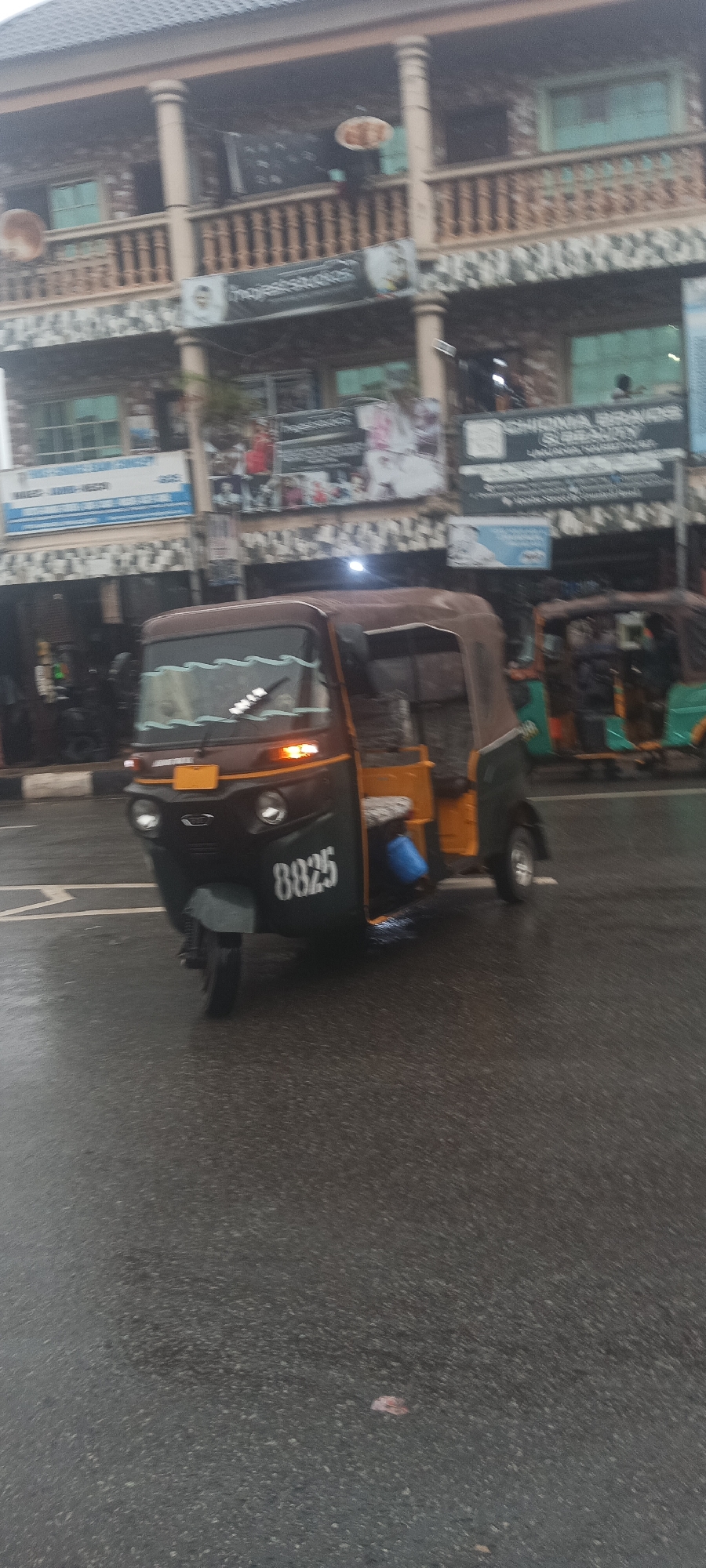The image depicts a motorized tricycle, commonly known as a "keke", in Umuahia, Nigeria. The keke is primarily dark brown and yellow with the number "8825" displayed on its front. The vehicle is moving on a wet asphalt road. In the background is a two-story building with a detailed balcony. The building appears to house multiple businesses, indicated by the signs above the storefronts. The overcast sky suggests recent or ongoing rain. The scene appears to be a common urban setting in Umuahia.