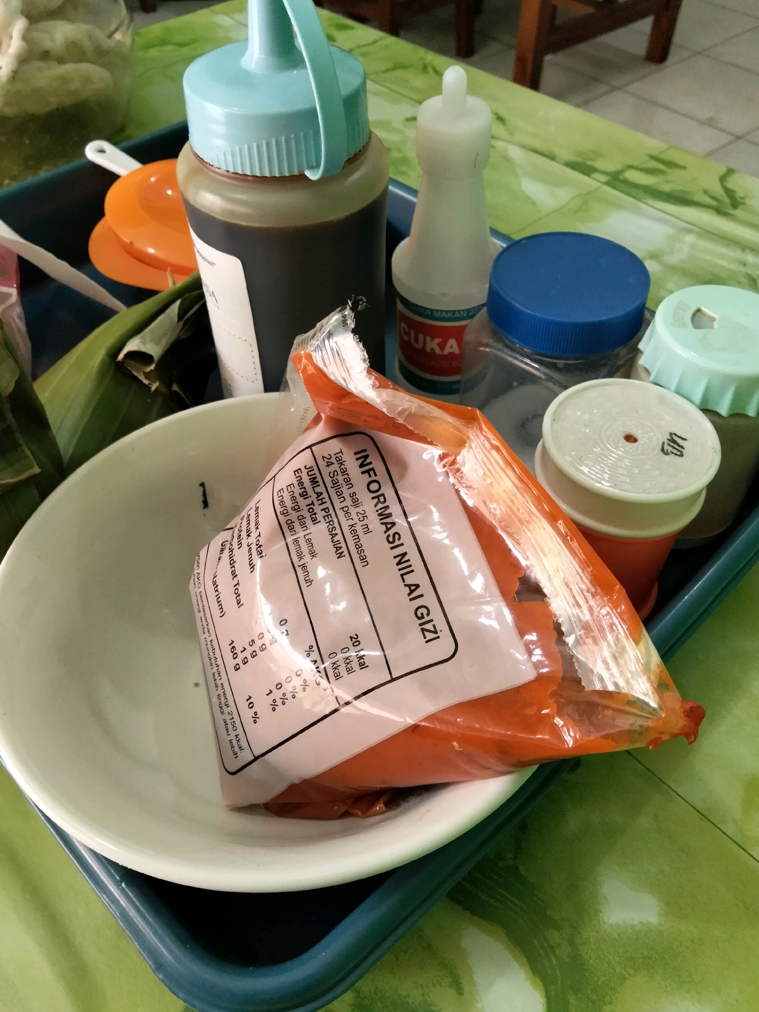 A close-up, eye-level shot captures a collection of condiment bottles and a packaged item within a blue plastic tray on a green, textured surface. The tray holds a large dark brown bottle with a light blue cap and spout, a white bottle labeled "CUKA MAKAN," a clear plastic container with a blue lid, and a smaller white container with a green lid. A partially opened orange plastic package containing a white and pink label with nutritional information is placed in a white bowl. The label details serving size, total calories, and macronutrient information. The background shows a wooden chair and a tiled floor, suggesting an indoor setting like a restaurant or food stall. The overall lighting is bright, with some shadows cast by the objects, indicating daylight or artificial light. The location is identified as Panggang, Indonesia, likely a food establishment in that area.