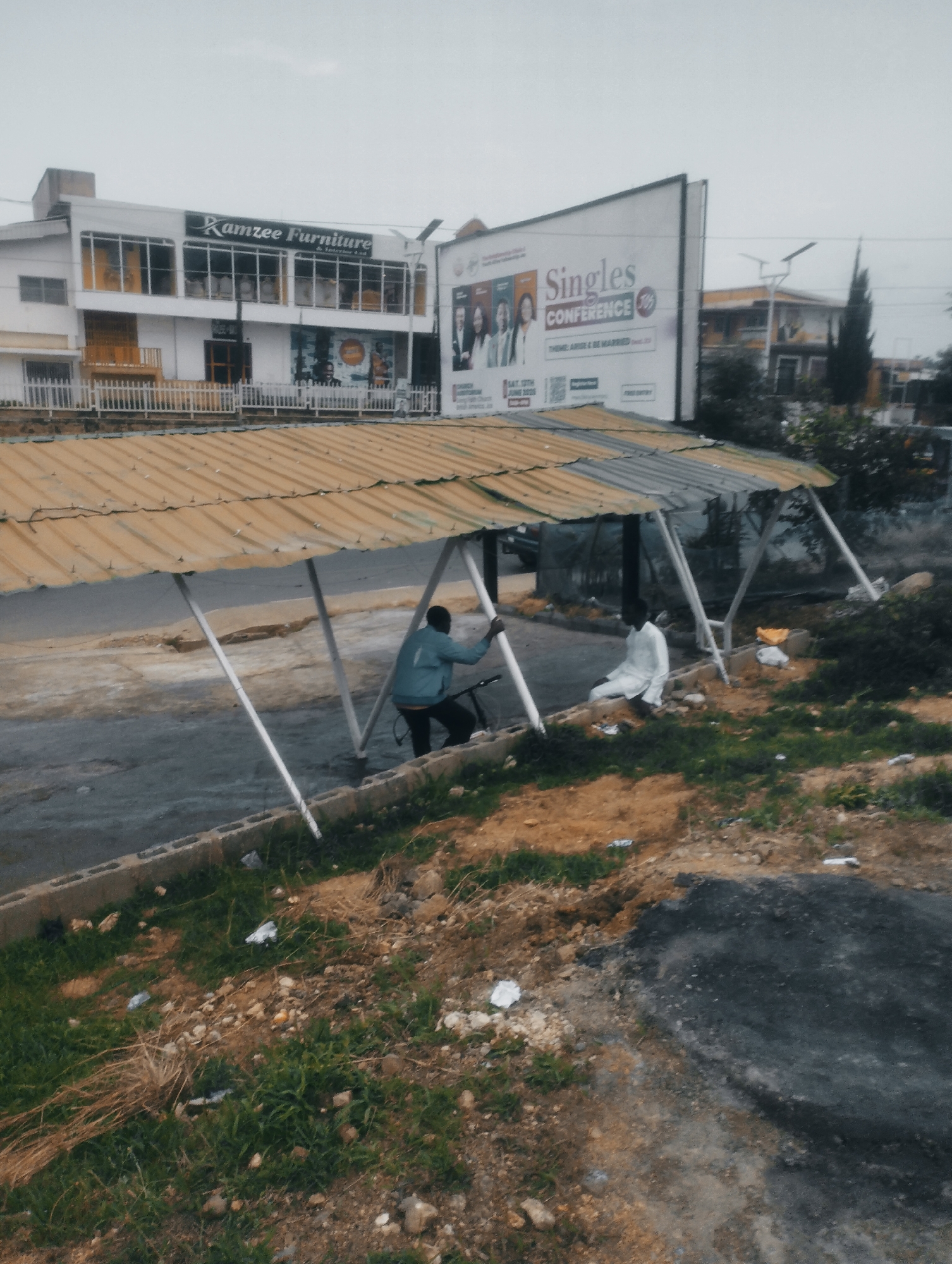 This is a medium shot, eye-level, outdoor photograph of a street scene in Jos, Nigeria. In the foreground, a yellow corrugated metal awning supported by white poles covers an area of rough asphalt and dirt. To the right, a person in a white garment sits on a raised dirt embankment. To the left, another person in a blue jacket stands next to a bicycle. In the background, a white building with "Ramzee Furniture & Interior" signage is visible. To the right of the building, a large billboard for a "Singles Conference" is mounted. The sky is overcast. The overall scene appears somewhat neglected, with scattered debris on the ground.