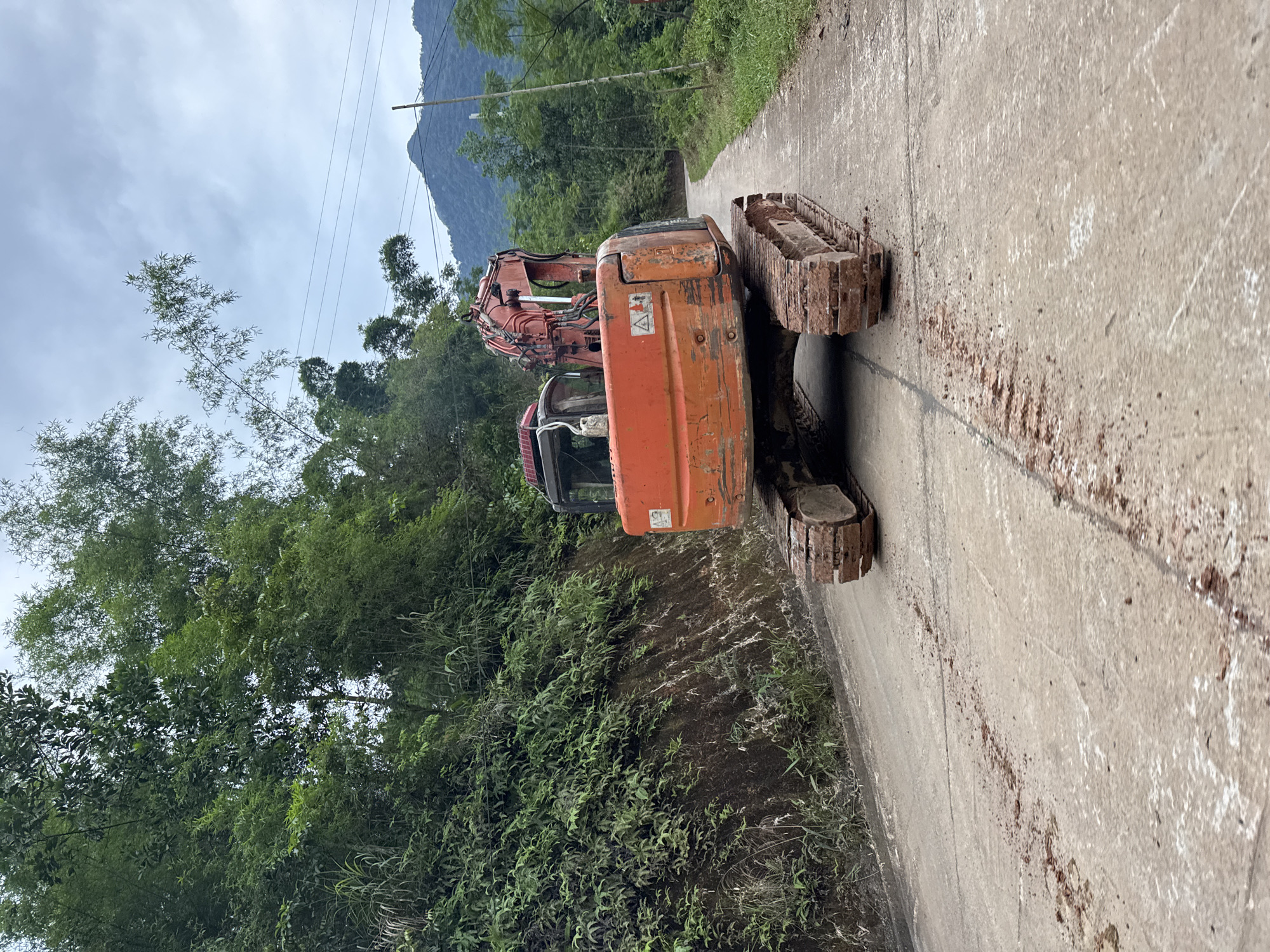 An orange excavator is stationary on a paved road in Cổ Lũng Commune, Vietnam. The road is wet and shows signs of mud or dirt. Lush green vegetation covers the steep embankment to the left of the road. In the background, a winding road leads towards rolling hills and mountains under a cloudy sky. Power lines are visible in the distance. The excavator appears to be the primary subject of the image, positioned centrally in the frame, facing away from the viewer and slightly to the right.