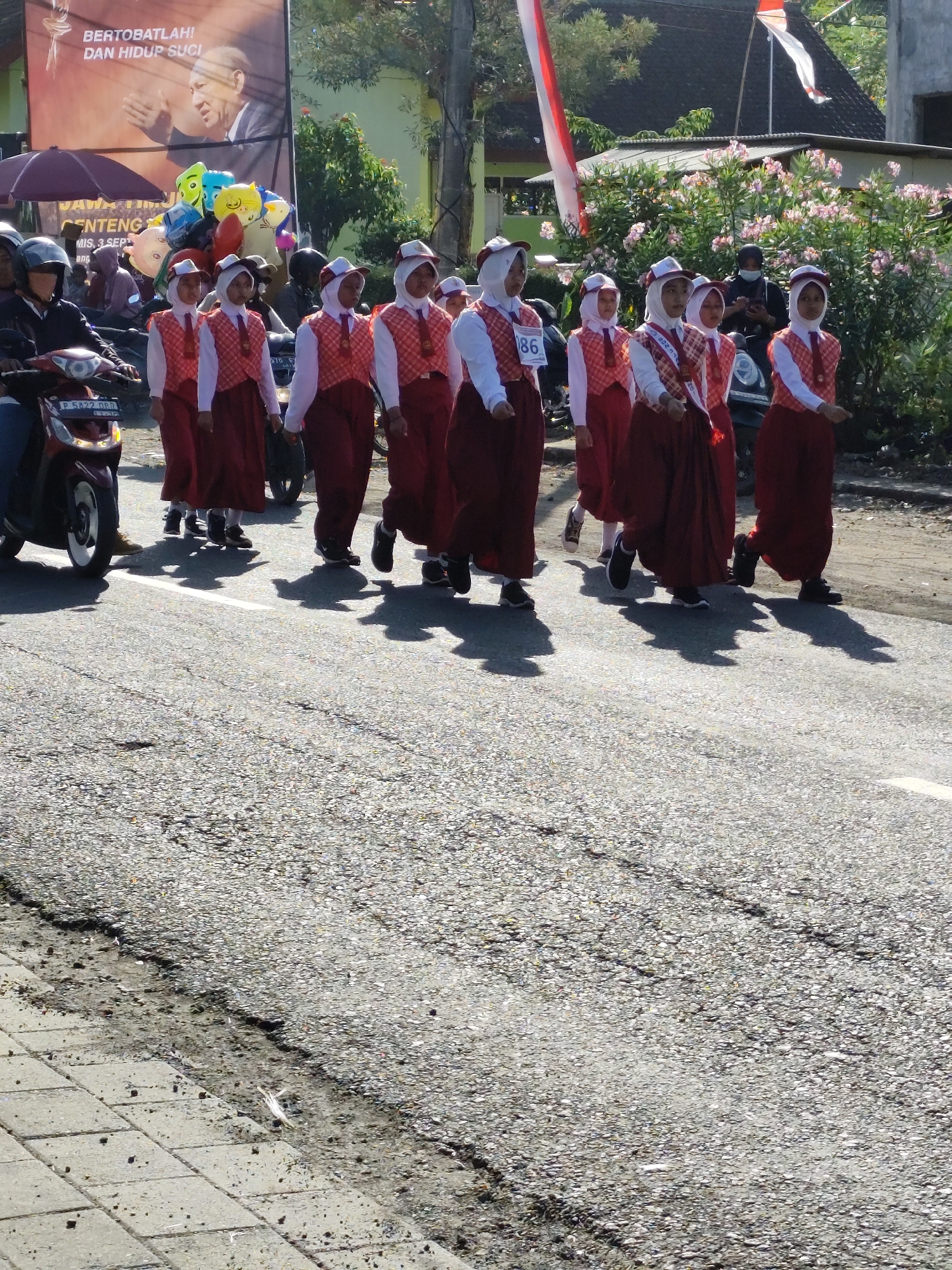 The image depicts a street scene in Tegalarum, Indonesia, likely during a parade or procession. In the foreground, a group of individuals, predominantly young women dressed in matching red and white uniforms with checkered vests, are walking along an asphalt road. They are wearing white headscarves and hats, and some have sashes or tags. Behind them, other participants and onlookers are visible, including a person on a scooter and others standing by the roadside. A large banner with text and an image of a man is displayed prominently on the left side of the image, along with colorful balloons. The environment appears to be a sunny day with trees and buildings in the background. The road is paved with asphalt and has a sidewalk made of concrete tiles on the left. The overall impression is of a community event taking place outdoors.