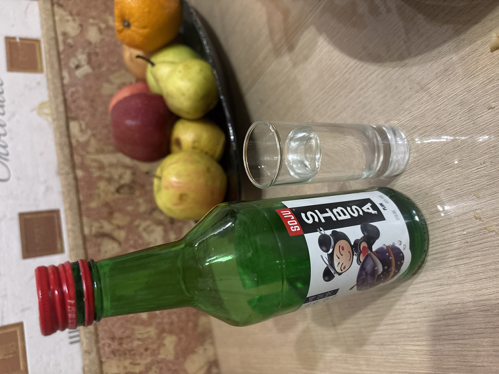 The image shows a still life scene. A green glass bottle with a red cap and a label that reads "SOJU" and "BTS" is laying horizontally on a wooden surface. Next to the bottle is a clear glass filled with a clear liquid, likely water. Adjacent to these items is a black bowl containing various fruits, including an orange, apples, and pears. The scene is set indoors on a table with a patterned table cloth. There is no evidence of the location being in Krasnoyarsk, Russia.