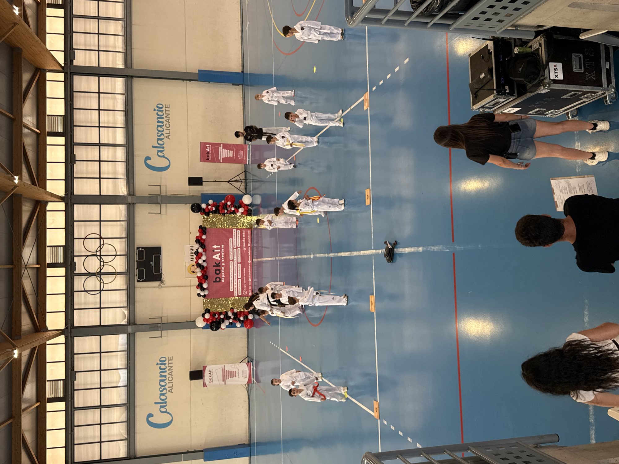 This image depicts a gymnasium interior, likely in a school or sports complex in Alicante, Spain, as suggested by the prominent "Calasancio Alicante" signage. A group of children wearing martial arts uniforms are lined up on the blue gymnasium floor. An adult in a black gi is positioned among them. Decorations, including balloons and a "bak Ait deporte y aventura" banner, indicate an event or celebration. Several adults are observing the scene. Some stage lighting and sound equipment are visible in the top-right corner.