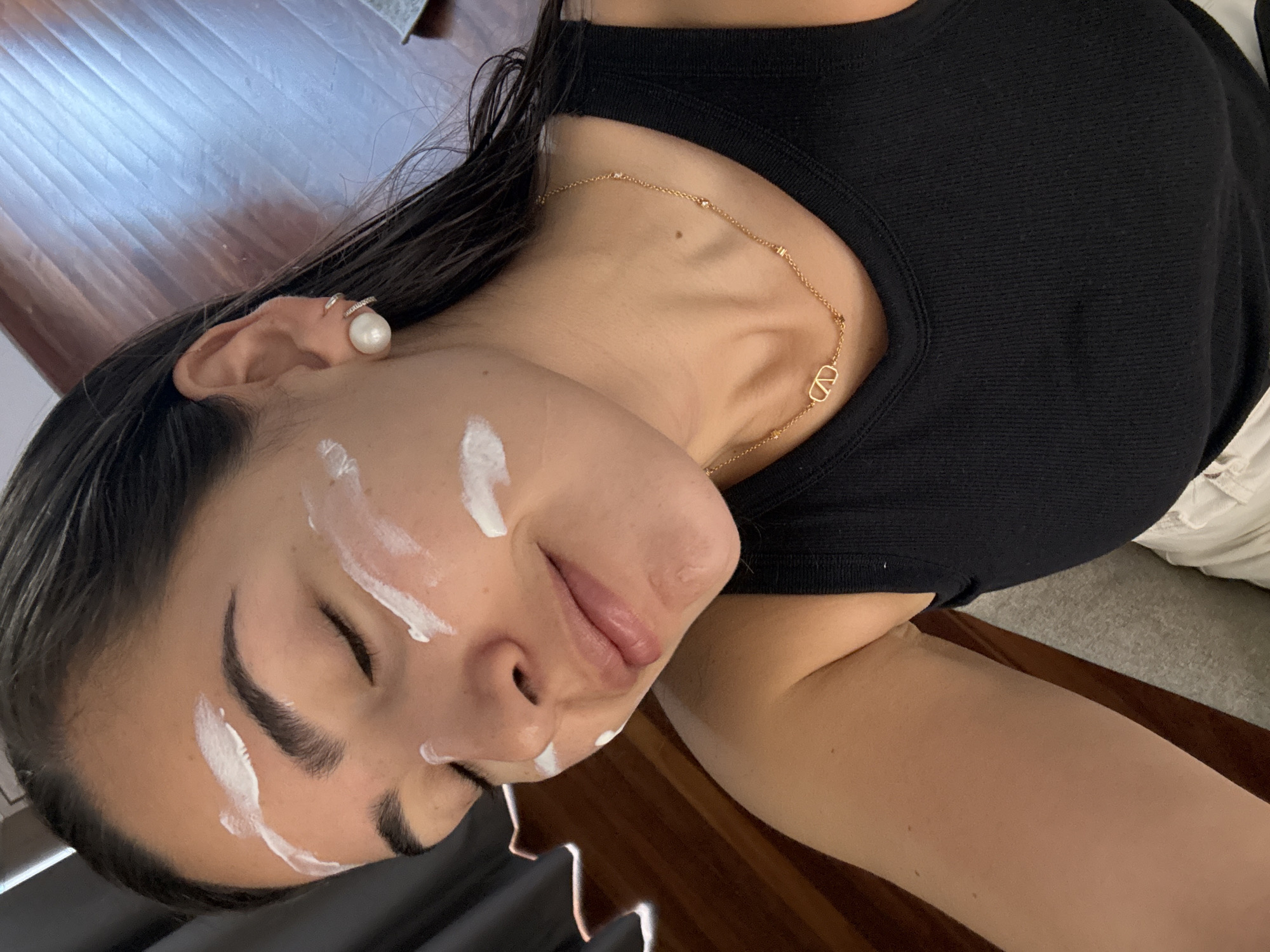 A young woman is reclining on a wooden surface. Her face has spots of white cream on the forehead, cheeks, and above the upper lip. She is wearing a black top and a gold necklace with a "V" pendant. A pearl earring is visible in her ear. The location in San Bernardino Tlaxcalancingo, Mexico, cannot be verified from the image.