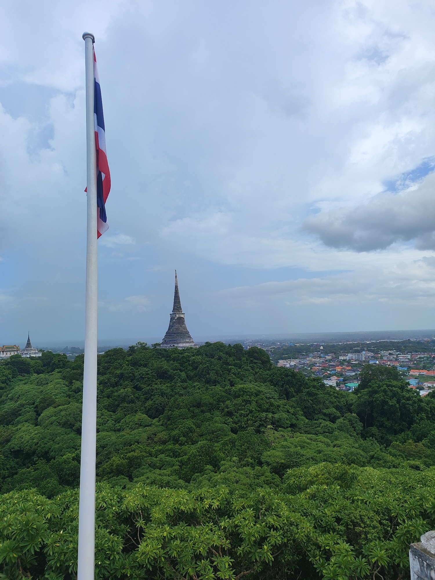 A panoramic view of Phetchaburi, Thailand, is captured from a high vantage point. In the foreground, a tall, white flagpole stands to the left, bearing the Thai flag, which is partially unfurled and shows its red, white, and blue stripes. The flag is positioned against a cloudy sky.

Dominating the midground is a lush, green forest covering rolling hills. Perched atop one of these hills, slightly to the right of center, is a distinctive, conical temple or stupa, its tiered structure visible against the hazy horizon. Further to the left, another similar architectural structure, appearing as a white building with a spire, is partially visible amongst the trees.

The background reveals the cityscape of Phetchaburi, a dense collection of buildings with varied rooftops, stretching across the flatter terrain beyond the forested hills. The sky is overcast with a mix of light and darker clouds, suggesting a potentially humid or rainy day. The overall scene conveys a sense of elevation and overlooks a significant portion of the city and its surrounding natural and religious landmarks.