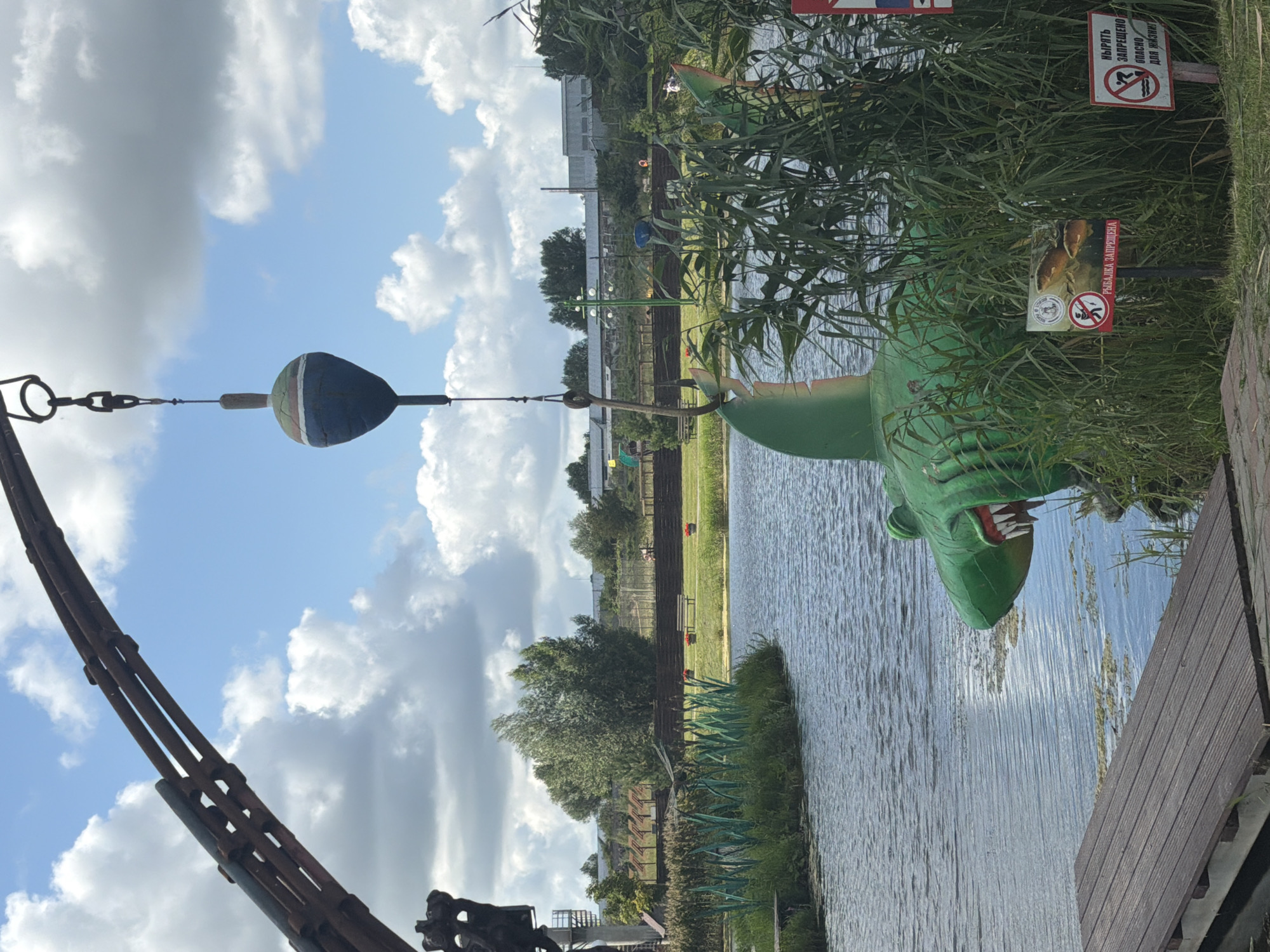 The image depicts a whimsical outdoor scene with a body of water, lush green reeds, and a cloudy sky. Dominating the foreground is a large, bright green shark sculpture with sharp white teeth, positioned as if emerging from the water. It is impaled on a large fishing hook suspended from a metal structure that arches into the frame from the top left. A fishing bobber hangs above the hook. To the left of the shark sculpture, a wooden dock extends into the water. In the background, trees and buildings are visible, suggesting a semi-rural or park-like setting. Several signs are placed near the shark sculpture. One sign, featuring an image of fish within a red circle and a diagonal line, has text that translates to "FISHING IS PROHIBITED." Another sign, with a similar prohibition symbol and an image of a person diving, reads "DIVING IS PROHIBITED. DANGEROUS TO LIFE." The overall impression is that of a decorative or recreational area, possibly an amusement park or a themed fishing spot. The location is identified as the city of Konstantinovka, Russia.