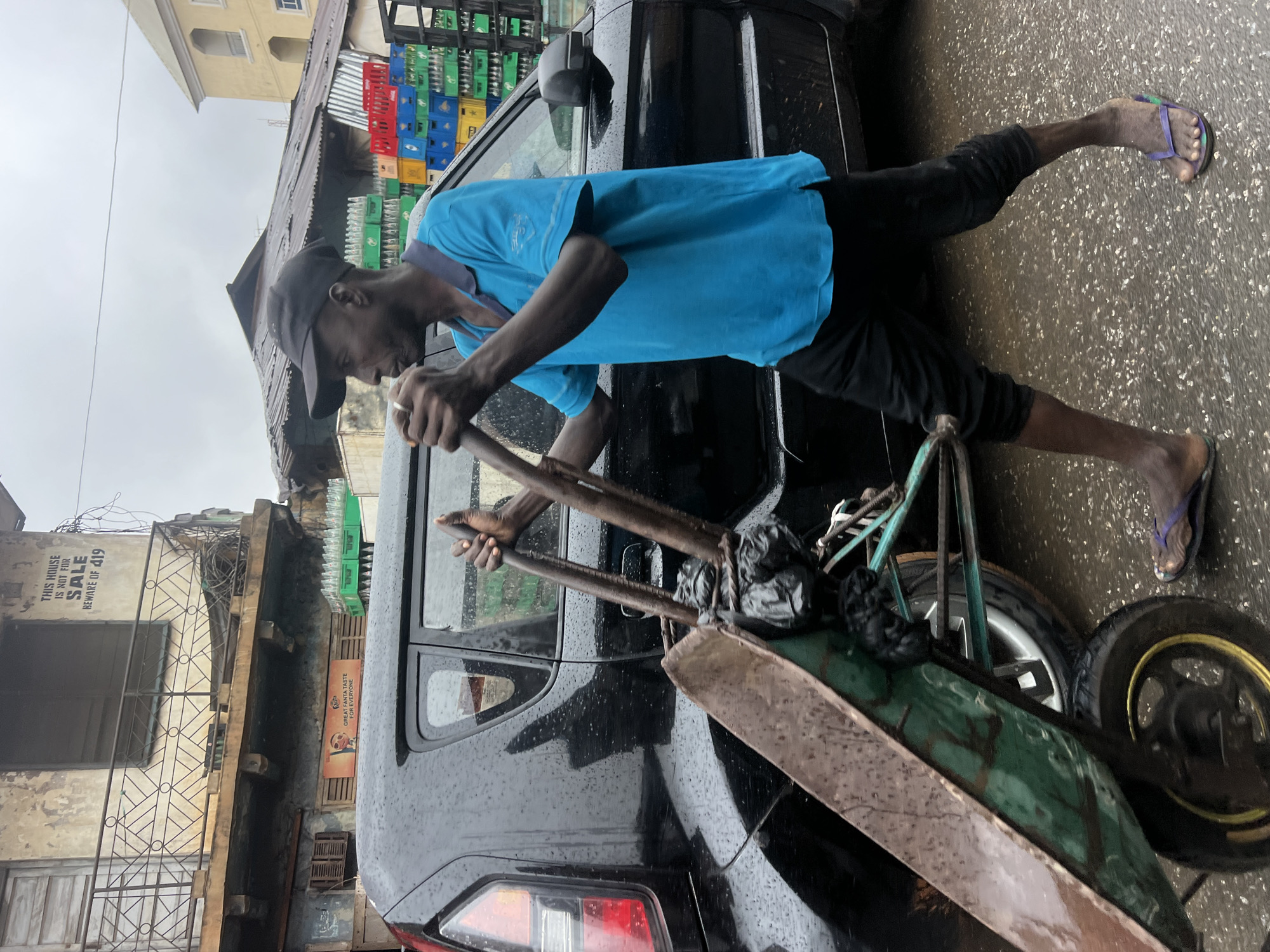 A man wearing a blue polo shirt, black pants, a black baseball cap, and flip-flops pushes a wheelbarrow next to a black car on a wet city street. The wheelbarrow is filled with black plastic bags and has a green metal basin. In the background, stacked crates of bottled drinks are visible, along with a weathered building with a balcony and a sign that reads "THIS HOUSE IS NOT FOR SALE BEWARE OF 419". The scene is likely in Onitsha, Nigeria, given the context of the "419" scam warning, which is a common reference to Nigerian fraud. The ground is wet, indicating recent rain, and water droplets are visible on the car's surface.