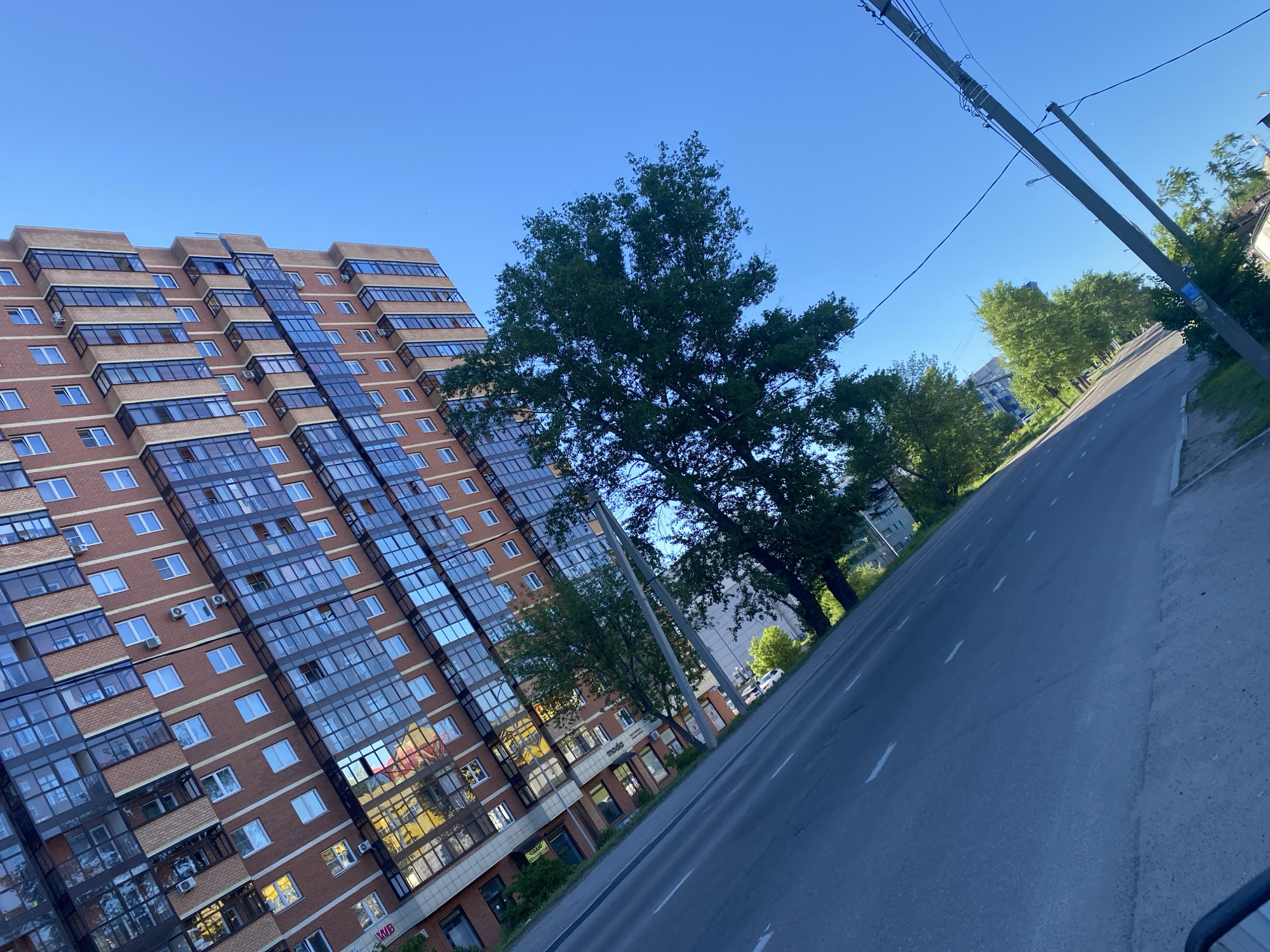 This is a daytime, eye-level, outdoor, wide-angle, high-resolution, full-shot photograph of a modern, multi-story apartment building in Irkutsk, Russia. The building, constructed with red brick and large, reflective glass windows, occupies the left side of the frame, appearing to recede into the distance at an angle. Many windows are visible, some with air conditioning units mounted outside. The building's facade has an angular design, with sections projecting outwards.
To the right of the apartment building is a paved road, also angled, with white dashed lines indicating lanes. A utility pole with wires runs along the right side of the road. Several mature trees with lush green foliage are interspersed between the building and the road, and also line the far side of the road, creating a semi-residential environment. The sky is clear and blue, indicating fair weather. There are no visible people or vehicles in the immediate scene. The overall impression is one of an urban residential area bathed in sunlight.