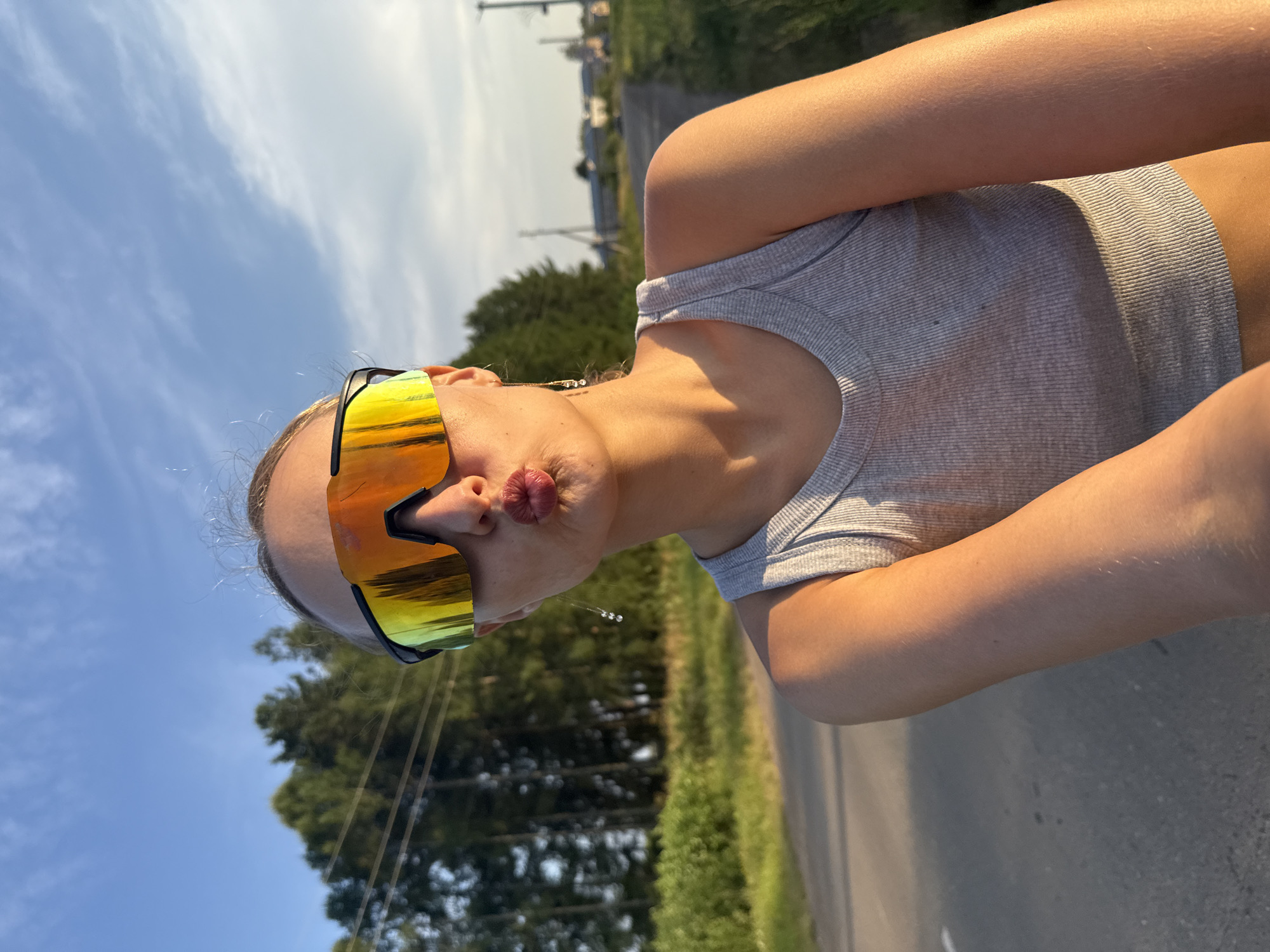 A medium shot captures a young woman posing for a selfie outdoors. She is wearing a grey ribbed tank top and oversized, reflective sunglasses with an orange and yellow tint. Her lips are puckered, and she has a small earring with three beads hanging from her right ear. The background suggests a rural setting with a paved road, a grassy verge with wildflowers, and a line of pine trees. In the distance, a few houses and a utility pole are visible. The sky is a clear blue with some wispy clouds, and the setting sun casts a warm, golden light on the scene, particularly on the woman's arm and shoulder. The location is the village of Podshivalovo, Russia.
