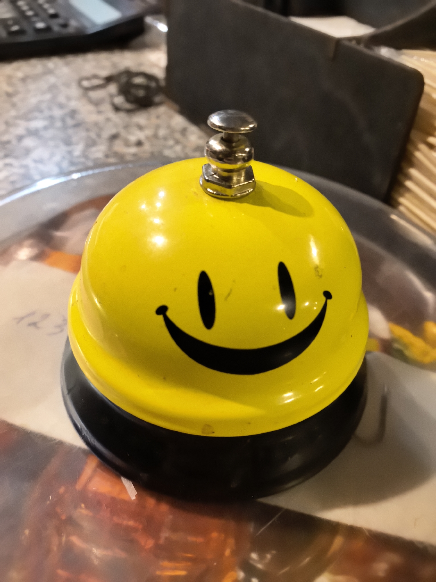A close-up shot depicts a yellow service bell with a black base and a simple smiley face on its surface. The bell is positioned on a reflective surface, possibly a tray, which displays indistinct reflections of surrounding objects and colors. In the background, out of focus, are a black calculator, a gray storage box, and wooden toothpicks. The location in Kamenka, Russia, cannot be determined from the visual information.