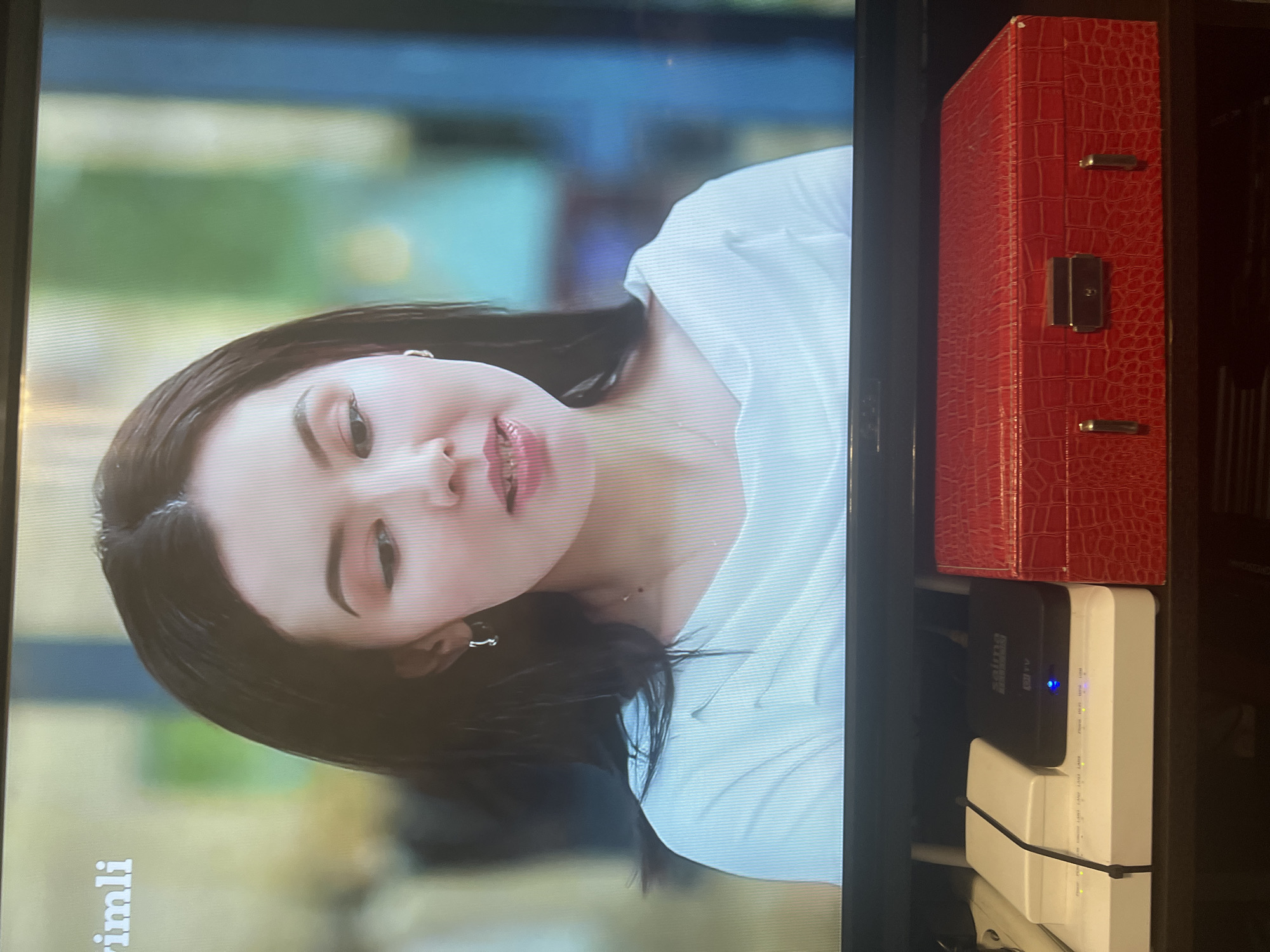 The image shows a television screen displaying a close-up of a young woman with dark hair and fair skin, wearing a white off-the-shoulder top and silver hoop earrings. She has on makeup, including eyeshadow and lipstick, and is looking downwards. Text is visible in the upper left corner of the screen, reading "imli". Below the television, there are electronic devices, including a white router, a black set-top box with the logo "saima", and a red textured box with a silver clasp. The television appears to be on a stand in a room. The location is identified as Osh City, Kyrgyzstan.