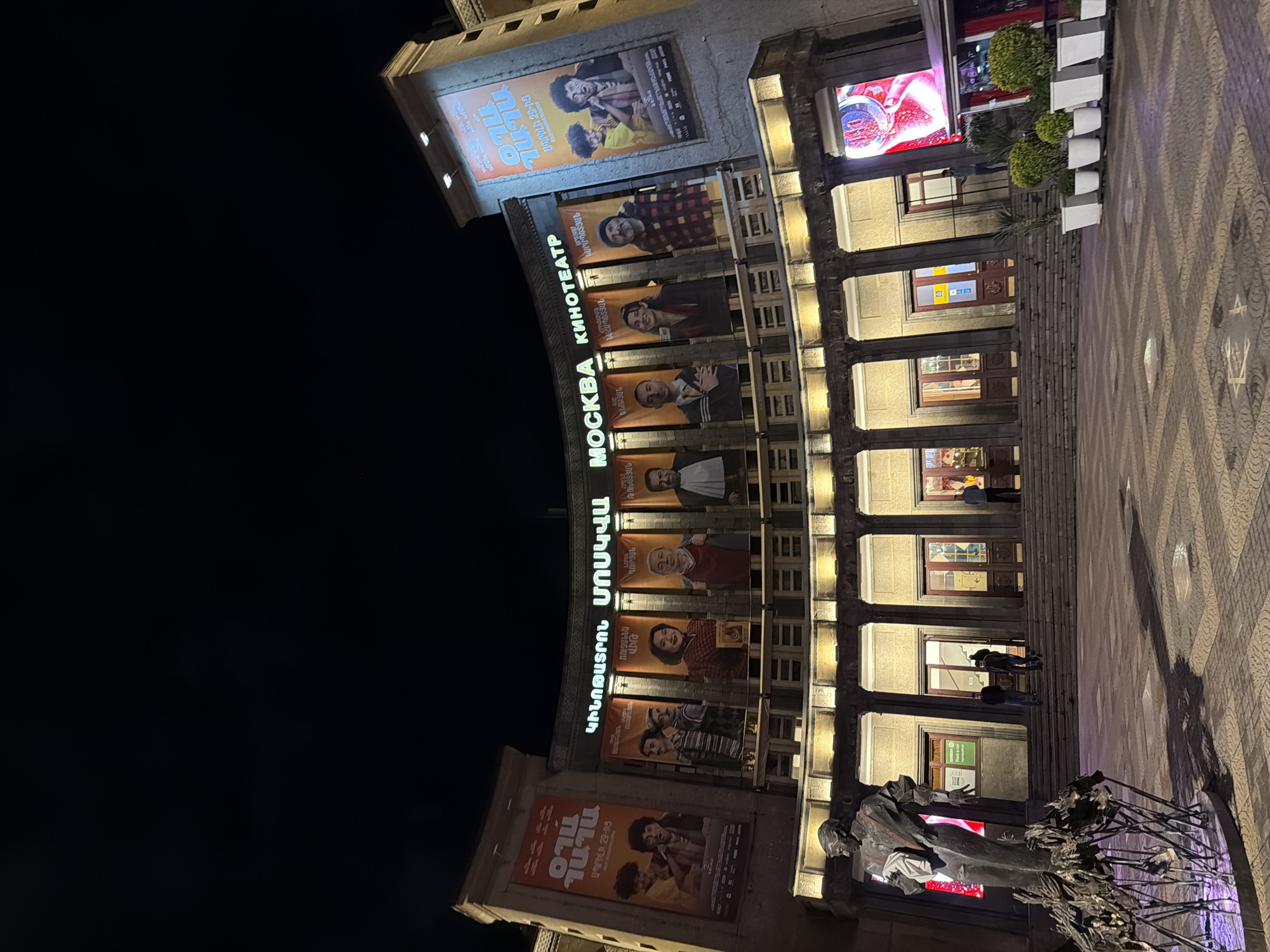 This vertical shot captures the exterior of a large building, identified as the "Moskva Cinema" or "Moscow Kinoteatr," at night in Yerevan, Armenia. The dark sky indicates nighttime, with the building's facade brightly illuminated by various lights and neon signs.
The cinema's name, "ՄՈՍԿՎԱ" (Moskva) in Armenian and "КИНОТЕАТР" (Kinoteatr) in Russian, is prominently displayed in large, glowing letters across the upper part of the building. Below these signs, the facade is adorned with numerous movie posters featuring diverse individuals and Armenian text, likely names of actors such as "ԿԱՐԱՊԵՏՅԱՆ," "ՌՈՒԶԱՆՆԱ," "ԴՈՒՐՅԱՆ," "ԹՈԽԱՏՅԱՆ," "ԿՆՈՋՅԱՆ," and "ՆԱՀԱՊԵՏՅԱՆ." Other texts like "TILO," "OIMIN," and "ԱՊՐԻԼԻ 29-ԻՑ" (from April 29th) suggest film titles and release dates.
The ground floor features a series of arched entrances and windows, supported by columns, creating a covered walkway. Warm light emanates from inside the building, illuminating the entrance area. Several people can be seen walking or standing near the entrances, suggesting active cinema operations or casual evening strolls. A sign for "Rent a car" is visible in one of the lower windows.
To the right, a patterned stone pavement extends across the foreground, leading up to the building. White planters with green shrubs are neatly arranged along the right side of the pavement. A large LED screen on the upper right displays vibrant, glowing content, possibly an advertisement. In the lower right corner, a dark-colored statue of a person, possibly an artist or historical figure, stands next to an abstract, dark metal sculpture, adding to the cultural ambiance of the scene. The overall impression is of a lively urban evening.