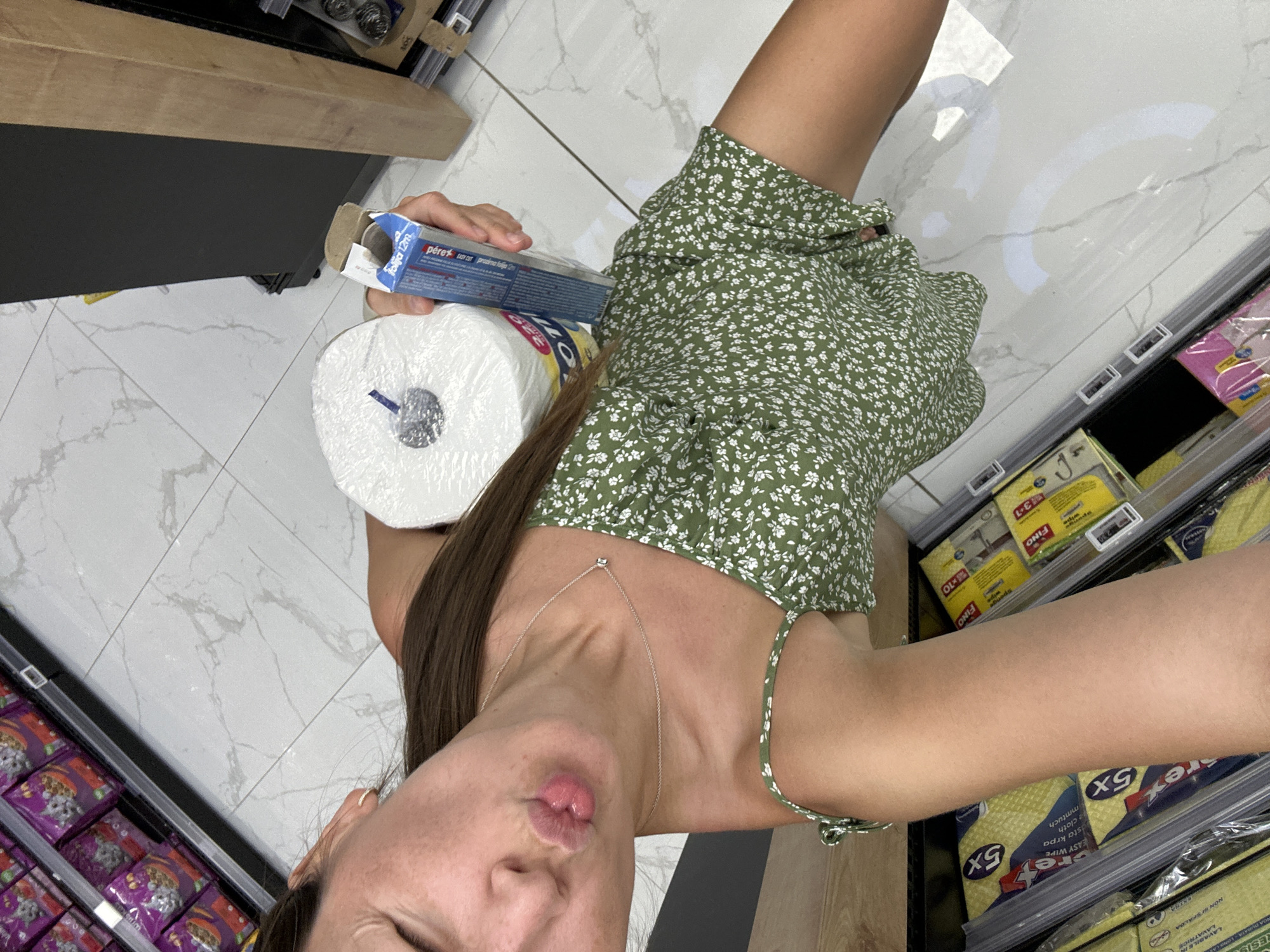 A young woman with dark hair is taking a selfie in a brightly lit supermarket aisle in Donji Pobori, Montenegro. She is wearing a green floral print dress and a delicate silver necklace. Her lips are pursed in a playful pout as she holds a large roll of paper towels and a box of aluminum foil. The supermarket shelves are stocked with various household items, including pet food, cleaning supplies, and paper products. The floor is tiled with a marble-like pattern, and natural light streams in, creating reflections on the polished surface. The overall scene captures a casual moment of shopping with a touch of lightheartedness.