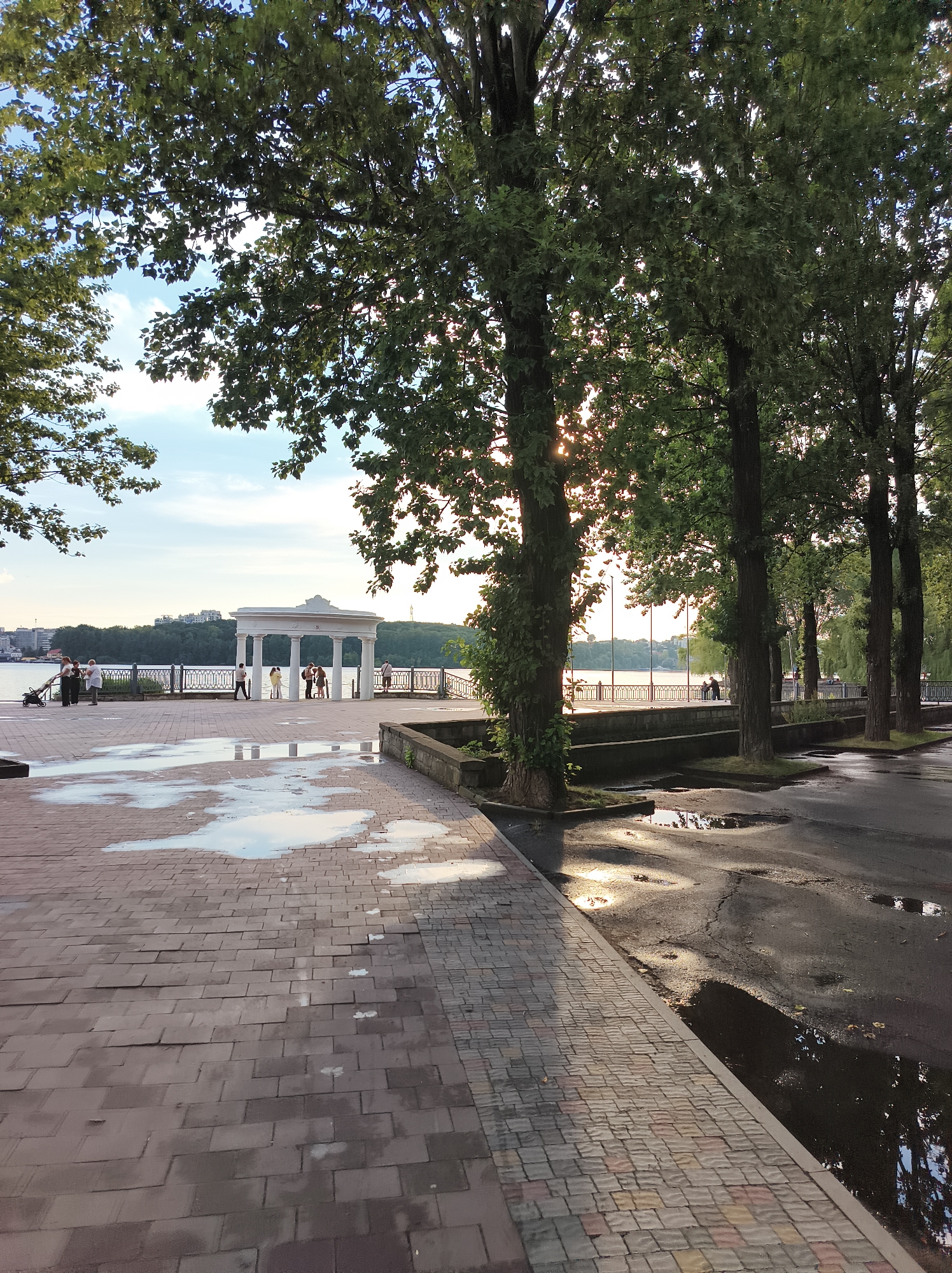 This image depicts a waterfront promenade in Ternopil, Ukraine, likely during the late afternoon or early evening, given the lighting. In the foreground, a brick-paved walkway is wet, with several large puddles reflecting the sky and surrounding trees. To the right, a dark asphalt road also shows reflective puddles. A row of tall trees with lush green foliage lines the walkway, their branches casting shadows.
Further along the promenade, near the water's edge, stands a white, classical-style rotunda with columns, serving as a focal point. Several people are visible on the promenade, some standing near the rotunda, others walking or standing by the water. A black metal railing separates the promenade from the body of water, which appears to be a lake or a wide river. In the distance, across the water, is a cityscape with buildings and a treeline along the horizon. The sky is partly cloudy with the sun visible through the trees, creating a soft, warm light.