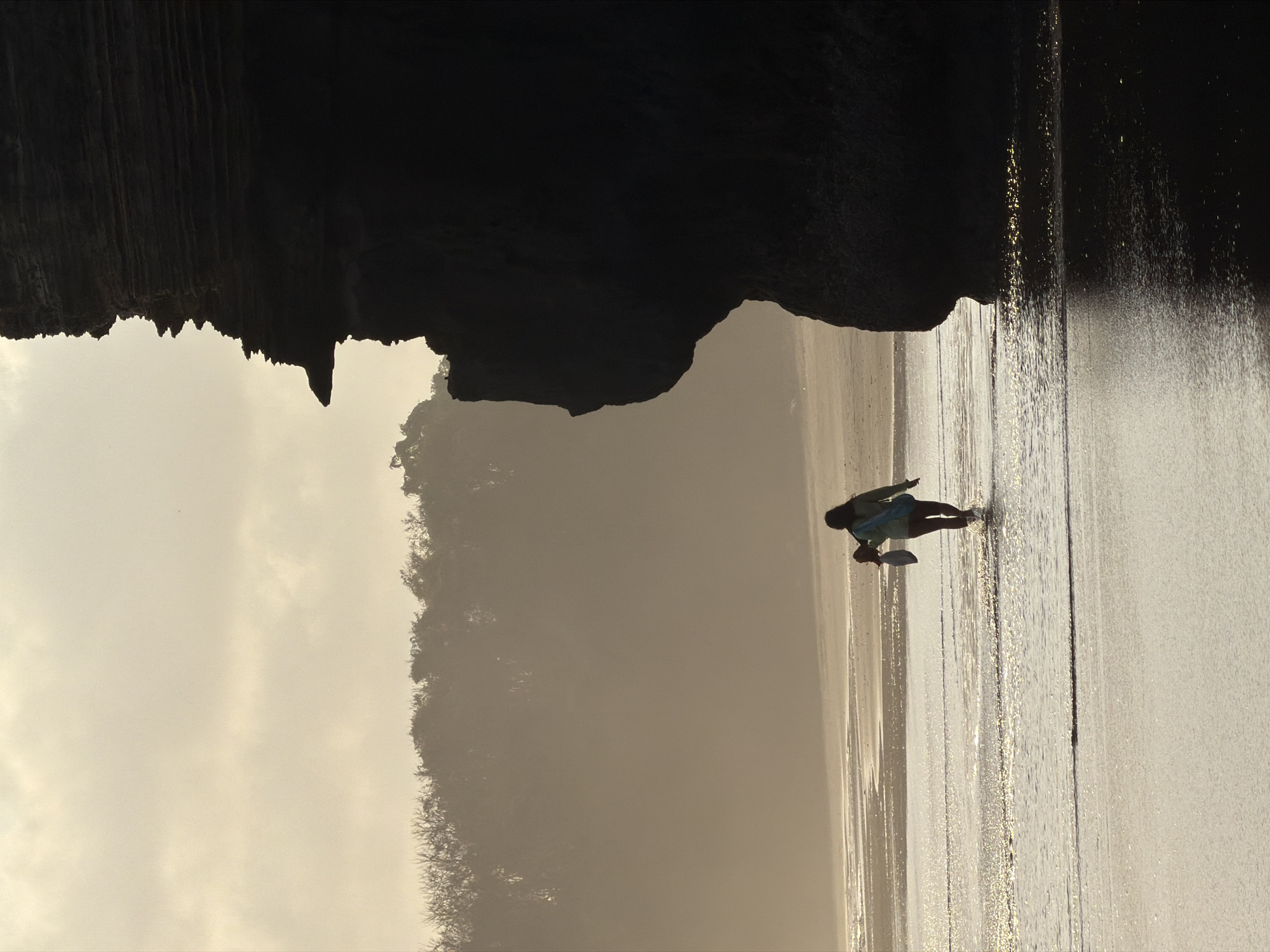 A solitary figure walks through shallow water on a beach in Selemadeg, Indonesia, carrying a bag over their shoulder and an object in their hand. The person is silhouetted against a hazy sky, with a large, dark rock formation dominating the right side of the frame. Trees are visible in the distance, partially obscured by mist. The water reflects the warm, diffused light of a setting or rising sun, creating shimmering patterns on the wet sand and shallow waves. The overall atmosphere is serene and slightly ethereal.