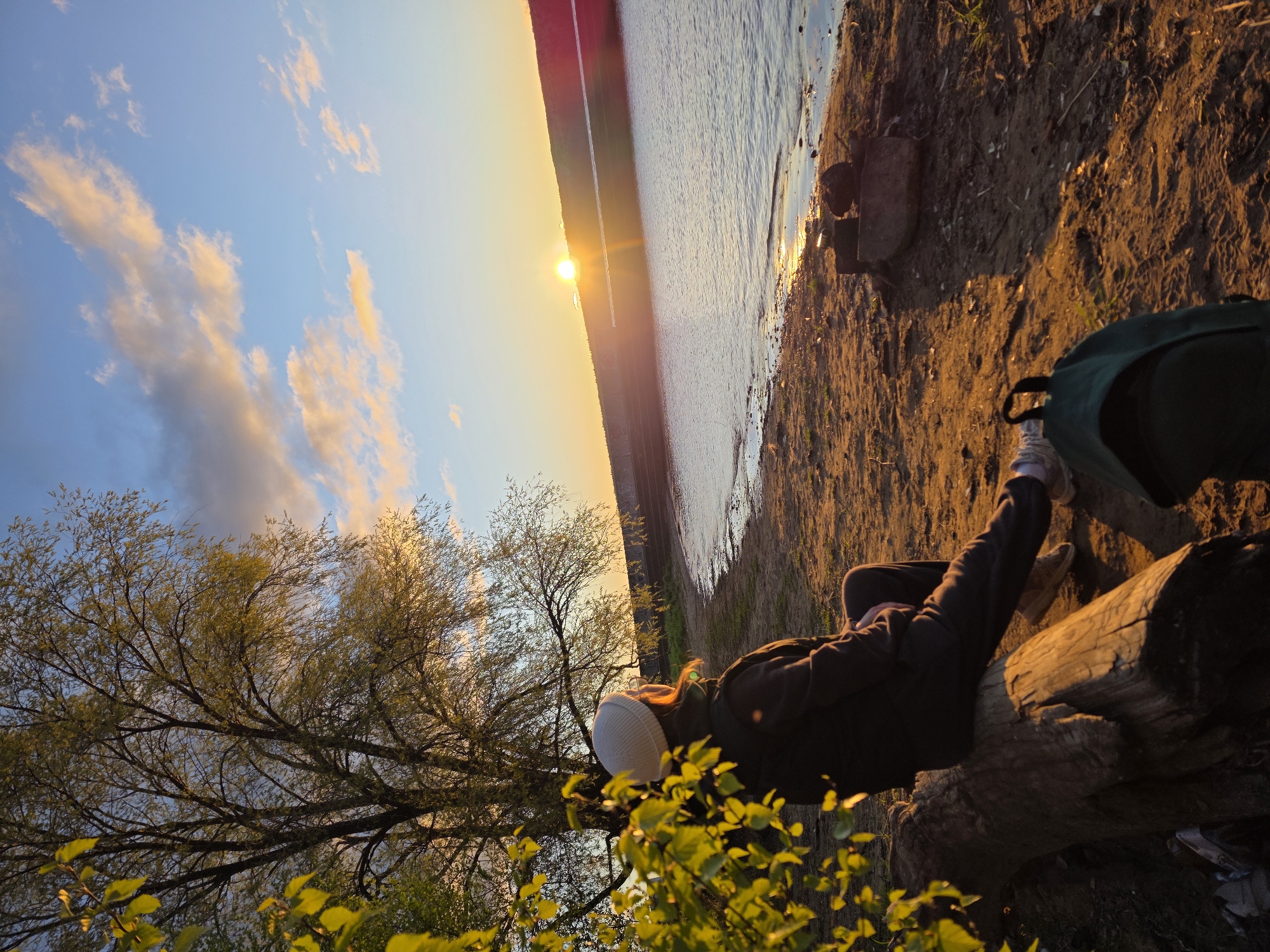 A person wearing a dark jacket and white cap rests on a log near the shore of the Kama River in Chaikovsky, Russia. The sun is setting over the river. The sky is blue with golden clouds. A tree with green leaves is in the foreground. A backpack rests on the ground near the log. The shore is sandy.