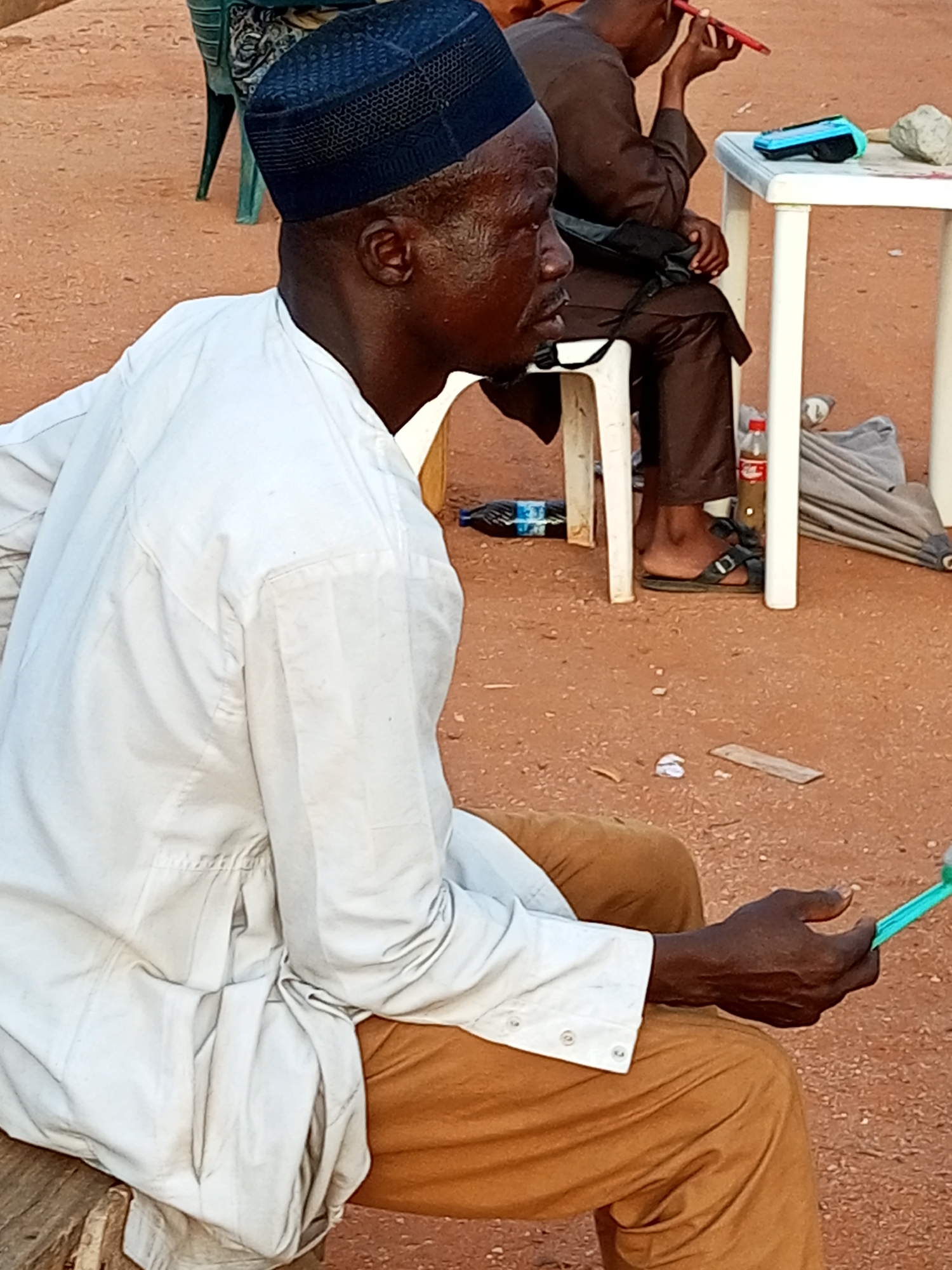 The image shows a medium shot of a Black man, appearing to be of middle age, sitting outdoors in what seems to be an informal gathering, possibly in Gombe, Nigeria. He is wearing a white long-sleeved traditional tunic and tan trousers, with a dark blue Kufi hat on his head. His skin is dark and appears moist. He is looking to his right and holding a bright green object, possibly a phone or a stylus, in his right hand. His left hand is resting on his lap.
In the background, another Black man in a brown outfit is partially visible, sitting on a white plastic chair and looking at something red he is holding to his eye. A white plastic table is to his right, with a turquoise electronic device and a rock on it. A bottle of Coca-Cola and a folded grey umbrella are also visible near the table. The ground is covered with reddish-brown soil and small stones. The scene suggests a casual, outdoor event.