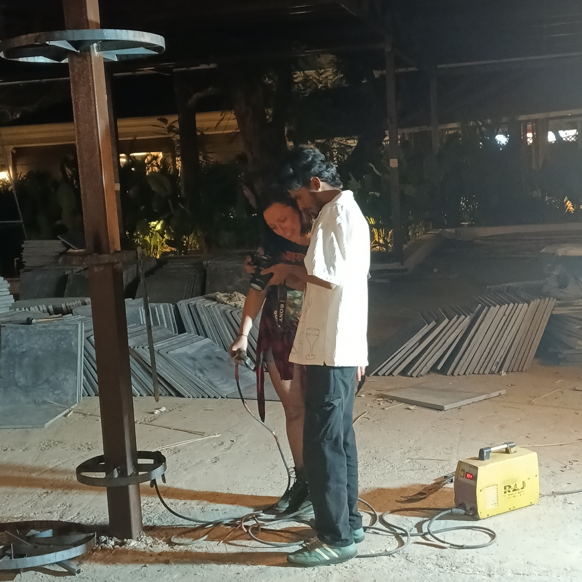 The image shows a man and a woman standing in an outdoor area under artificial lighting, potentially in Panaji, India. They are both looking at a digital camera, which the woman is holding, possibly reviewing photos. The man is wearing a white shirt with a wine glass design on it and dark trousers, while the woman is wearing a dark top with the Sony logo on the camera strap, and a red skirt or wrap.
The environment appears to be a construction or renovation site, with stacks of grey slabs of stone or concrete tile arranged in neat piles in the background. A metal structure, possibly a lighting fixture or construction support, stands to the left, with a welding machine on the ground beside it. The ground is sandy or dusty. The setting is outdoors and the presence of stone slabs indicates that it may be a construction site.