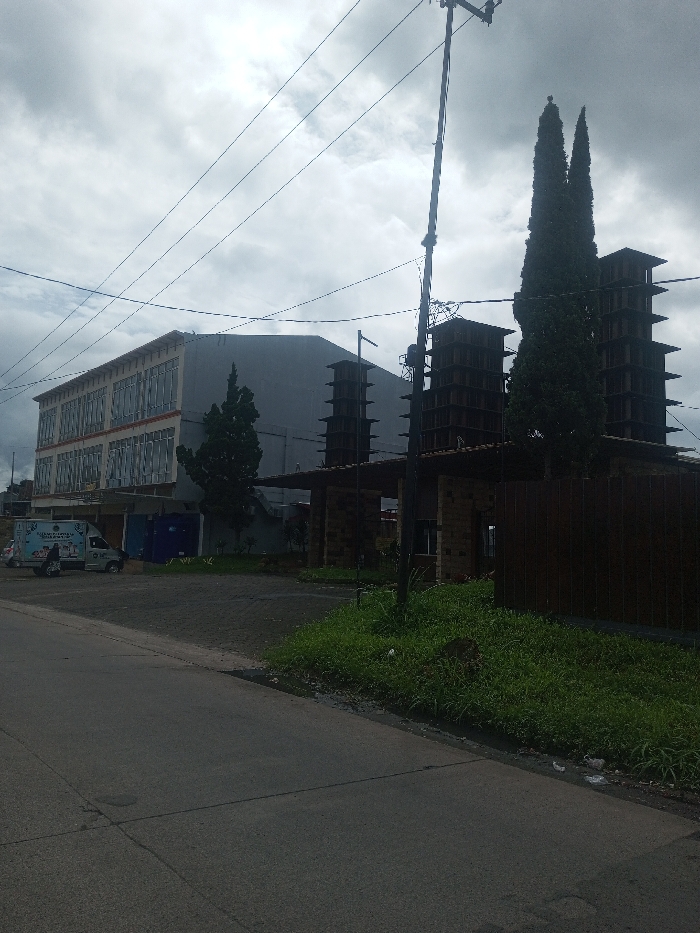 The image depicts an outdoor scene in Sukanagalih, Indonesia, under an overcast sky. In the foreground, an asphalt road runs left to right, bordered by a grassy verge on its right edge. To the left of the road, a two-story building with a light-colored facade and multiple rectangular windows is visible. A white cargo van displaying blue graphics and a dark scooter are parked in front of this building. Further right, a stone-clad entrance structure with a flat canopy leads to an area featuring three dark, multi-tiered tower-like structures. Two tall, slender conifer trees are positioned near these towers. A utility pole supporting multiple overhead power lines is visible in the mid-right portion of the frame. No human figures are present.