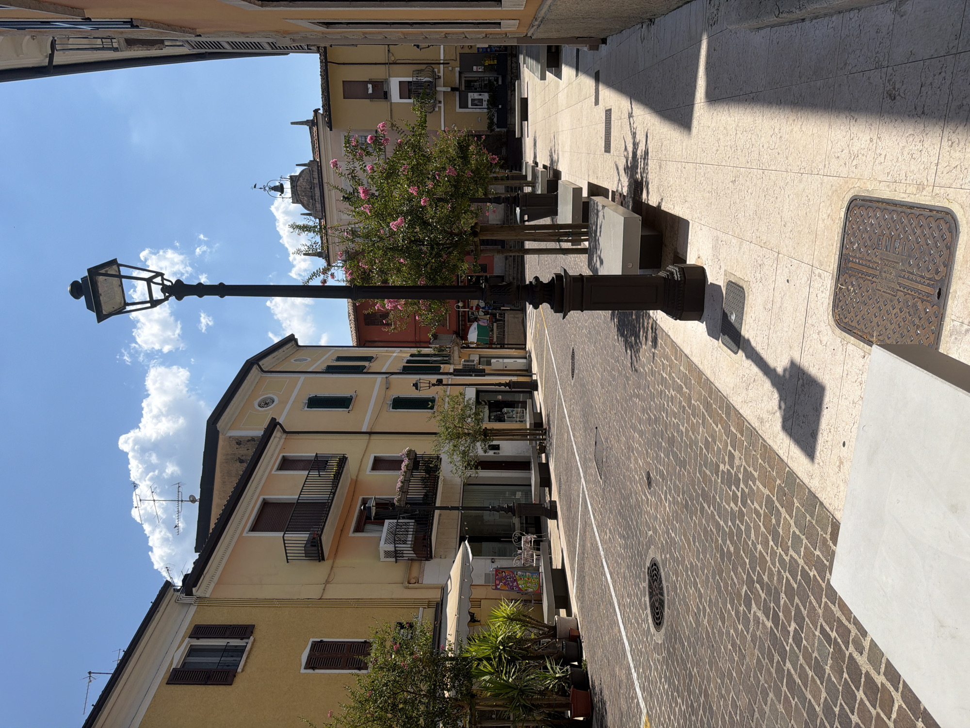 The image depicts a sunlit street in Padenghe sul Garda, Italy, with buildings lining both sides. The street is paved with cobblestones on the left and large light-colored stone tiles on the right, separated by white lines. Several lampposts are present, the most prominent being a tall black one in the foreground. Modern concrete benches are placed along the sidewalk on the right. Trees with pink blossoms are planted along the street, adding color to the scene. The buildings are mostly painted in warm yellow and cream tones, with some featuring balconies and shutters. The sky is bright blue with scattered white clouds. There are no people visible in the image. The overall impression is one of a quiet and pleasant urban environment.