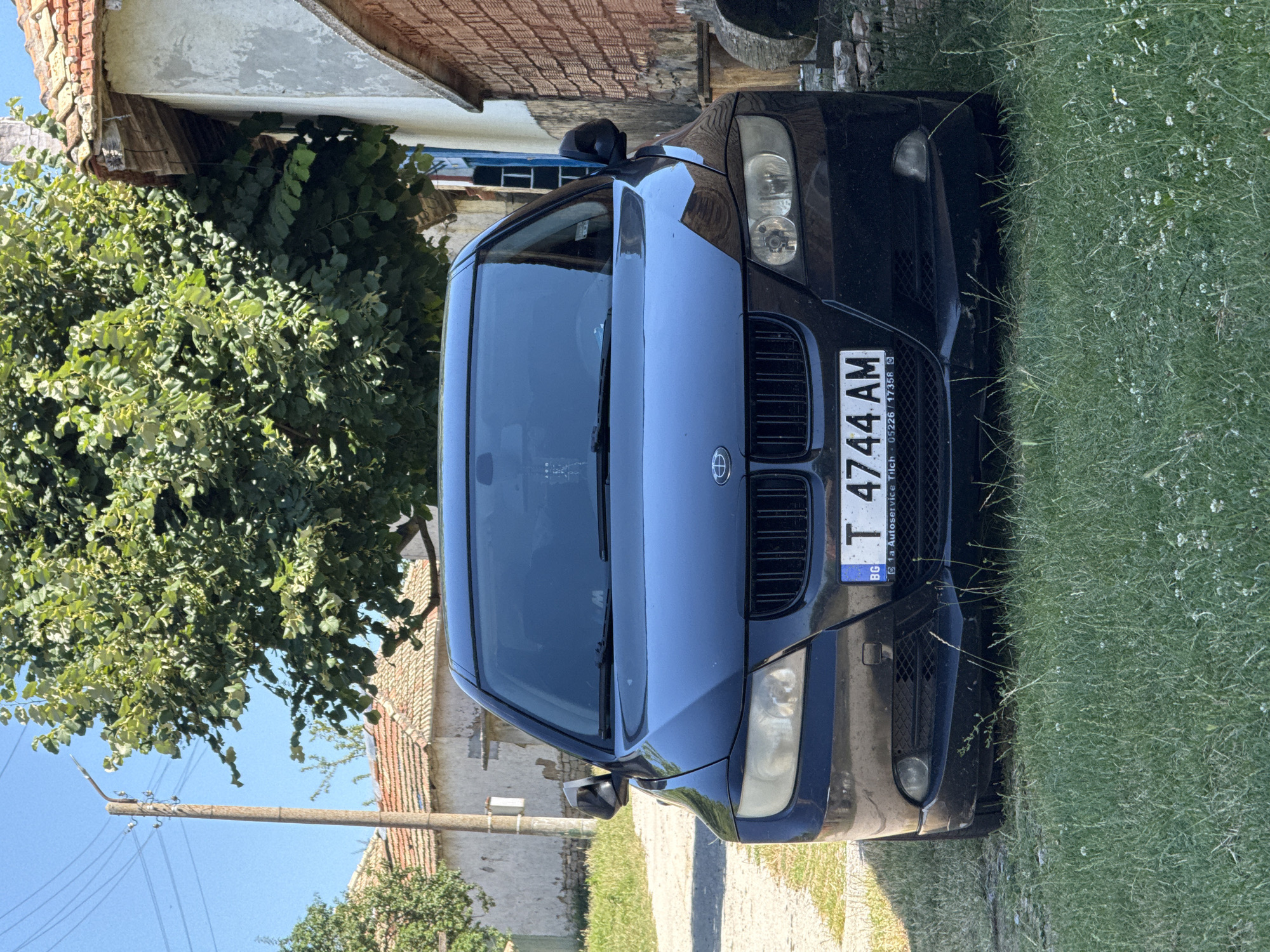 A black BMW sedan is parked on a grassy area in front of two weathered buildings, one with a red-tiled roof and the other made of brick. A large tree with green leaves partially shades the scene. A wooden utility pole with wires is visible to the left of the car. The car's license plate reads "T 4744 AM". The scene appears to be in Konop, Bulgaria.